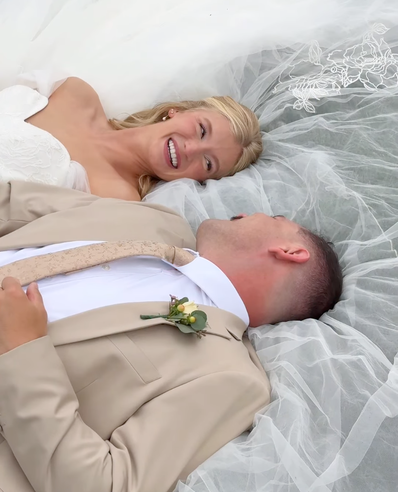 A bride and groom lie on the floor in wedding attire, smiling and gazing at each other, surrounded by white wedding decorations.