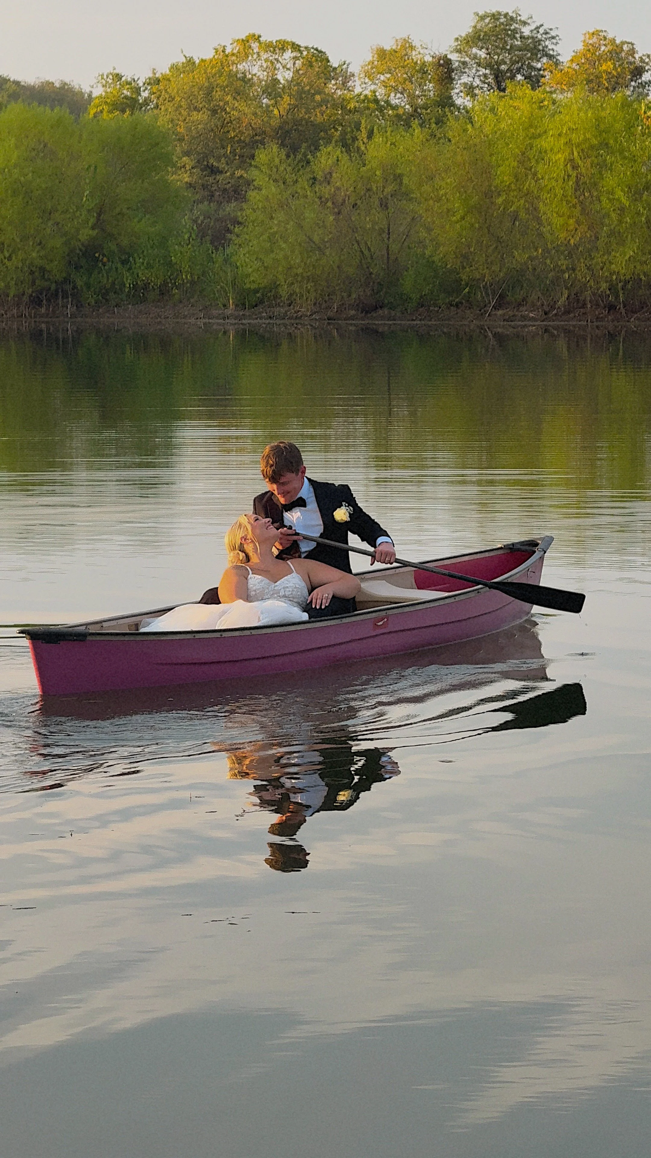 A couple dressed in wedding attire in a pink canoe on a calm river with green trees in the background.