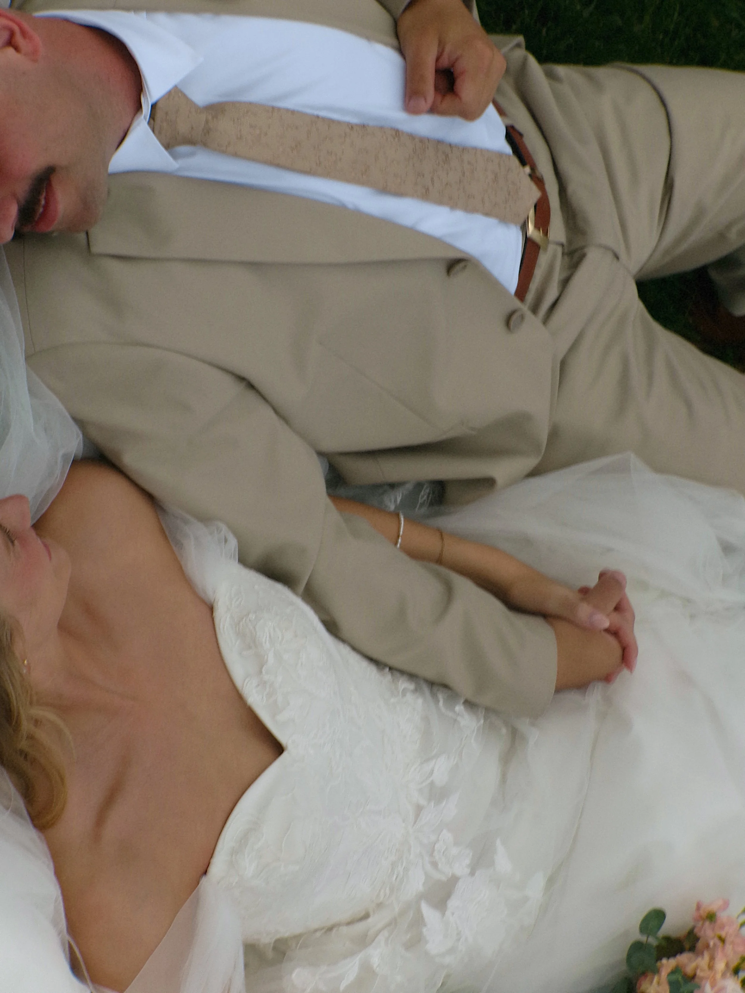 Close-up of a woman and a man lying down together, holding hands. The woman is wearing a white lace dress with an off-shoulder design, and the man is wearing a beige suit with a light pink tie and shirt. There are pink flowers and green leaves visible at the bottom right of the image.