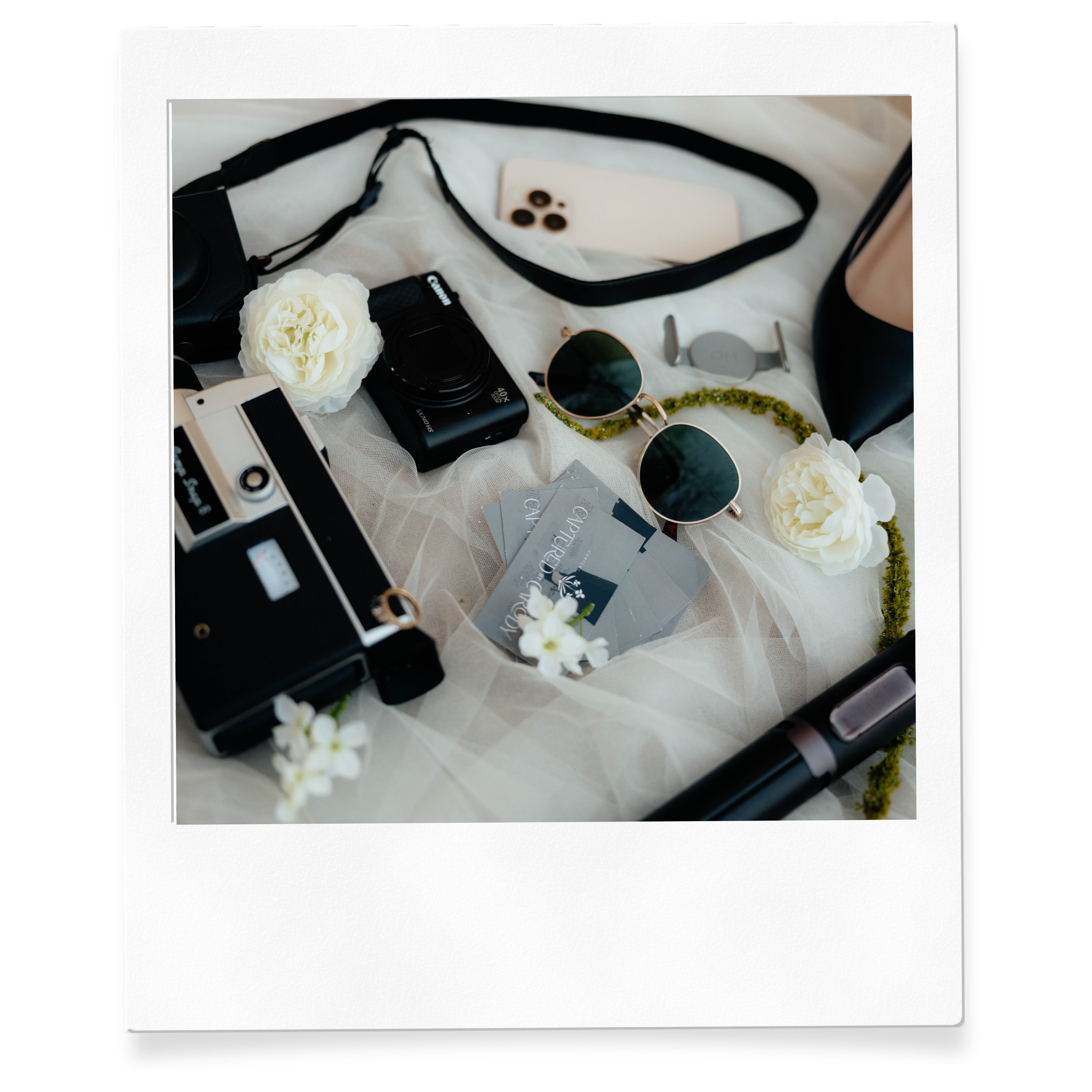 A flat lay of various items including a camera, sunglasses, a ring, a necklace, a white flower, a smartphone, and tags, placed on a white fabric surface.