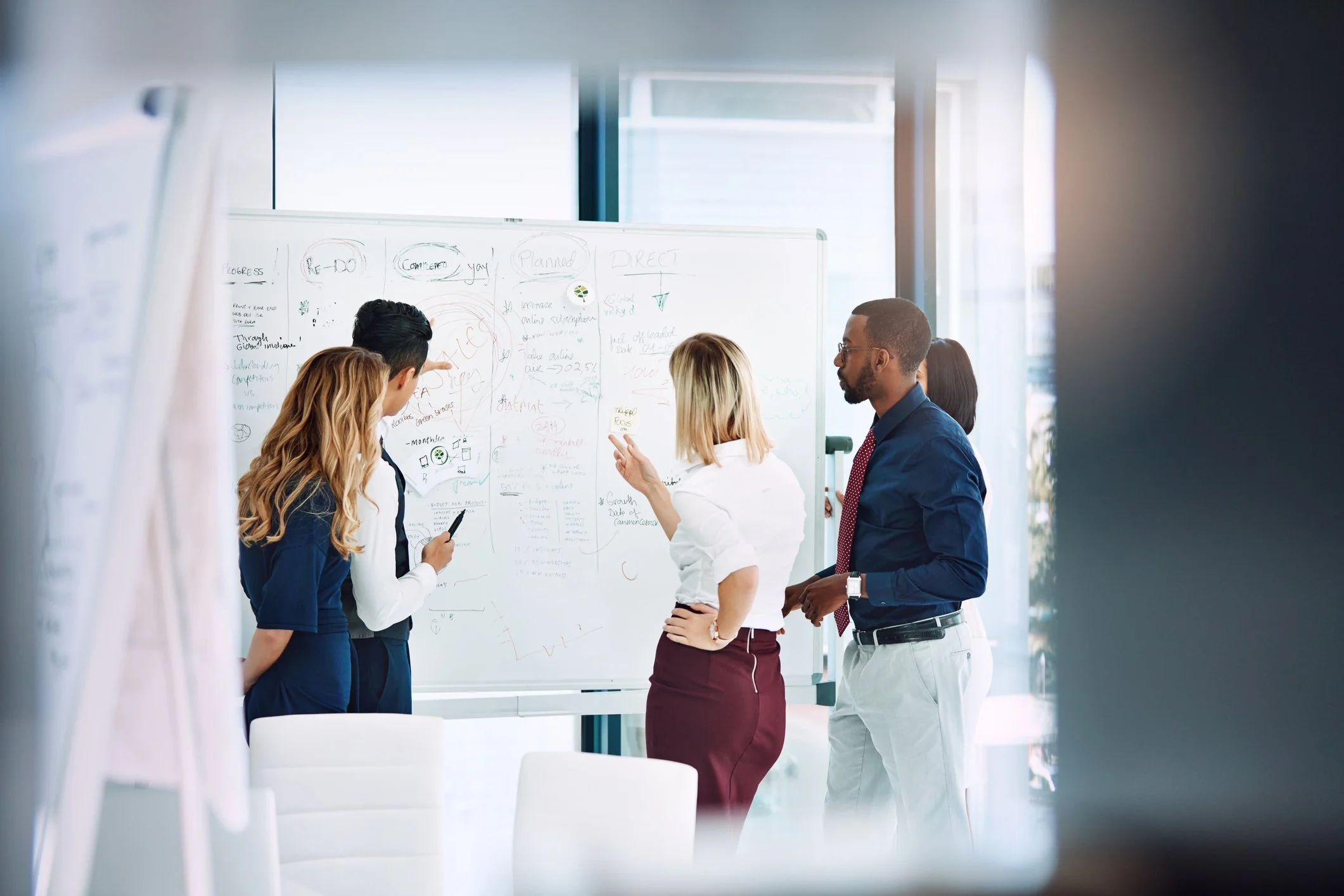 Group of five professionals discussing ideas around a whiteboard covered with notes and diagrams in a modern conference room.