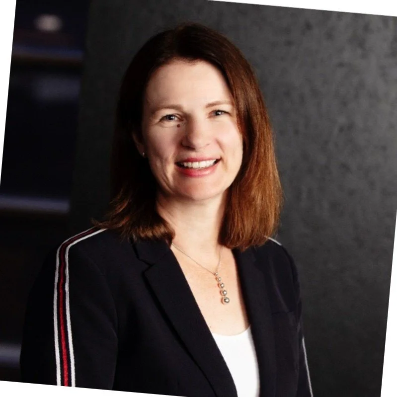 A woman with shoulder-length reddish-brown hair smiling, wearing a black blazer with white and red striped details on the sleeves, and a white top, standing against a dark gray background.
