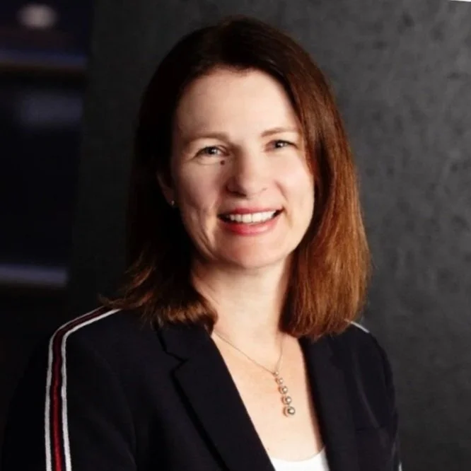 A woman with shoulder-length brown hair smiling at the camera, wearing a black blazer with stripes on the sleeves and a necklace, standing against a dark background.