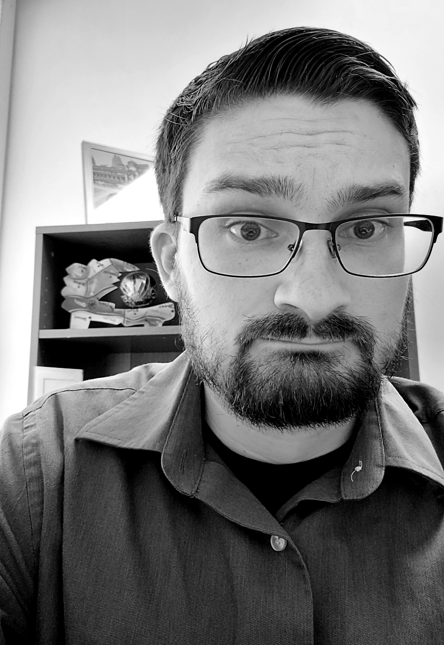 Black and white close-up photo of a man with glasses, a beard, and mustache, wearing a collared shirt, sitting in an office with a bookshelf and picture frame in the background.