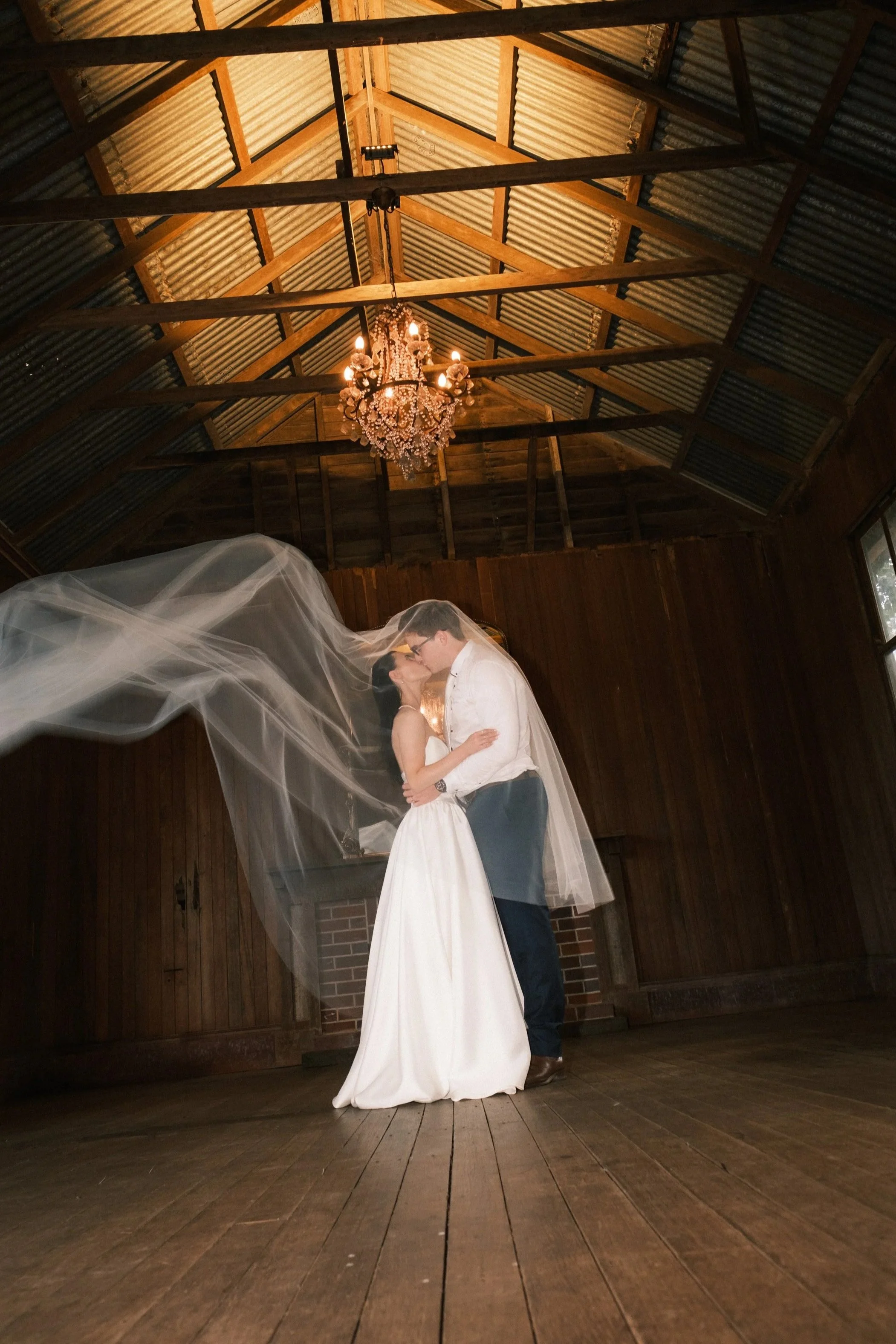 A bride and groom kissing in a rustic wooden venue with a chandelier hanging from a high ceiling, the bride wearing a white wedding gown and veil, the groom in a white shirt and dark pants, the bride's veil flowing in the air.