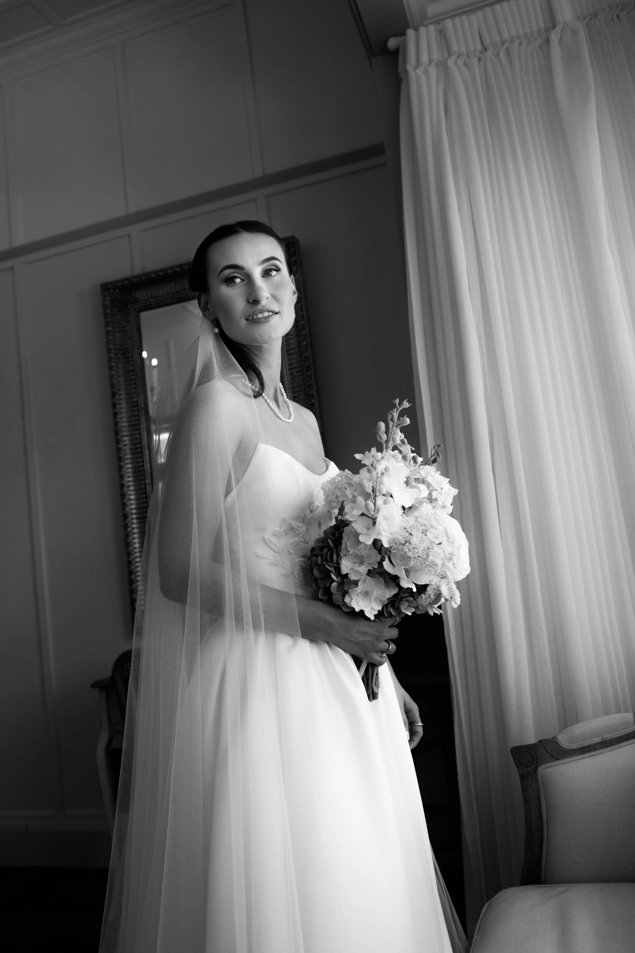 A black and white photo of a bride in a wedding dress holding a bouquet of flowers, standing indoors near a window with curtains.