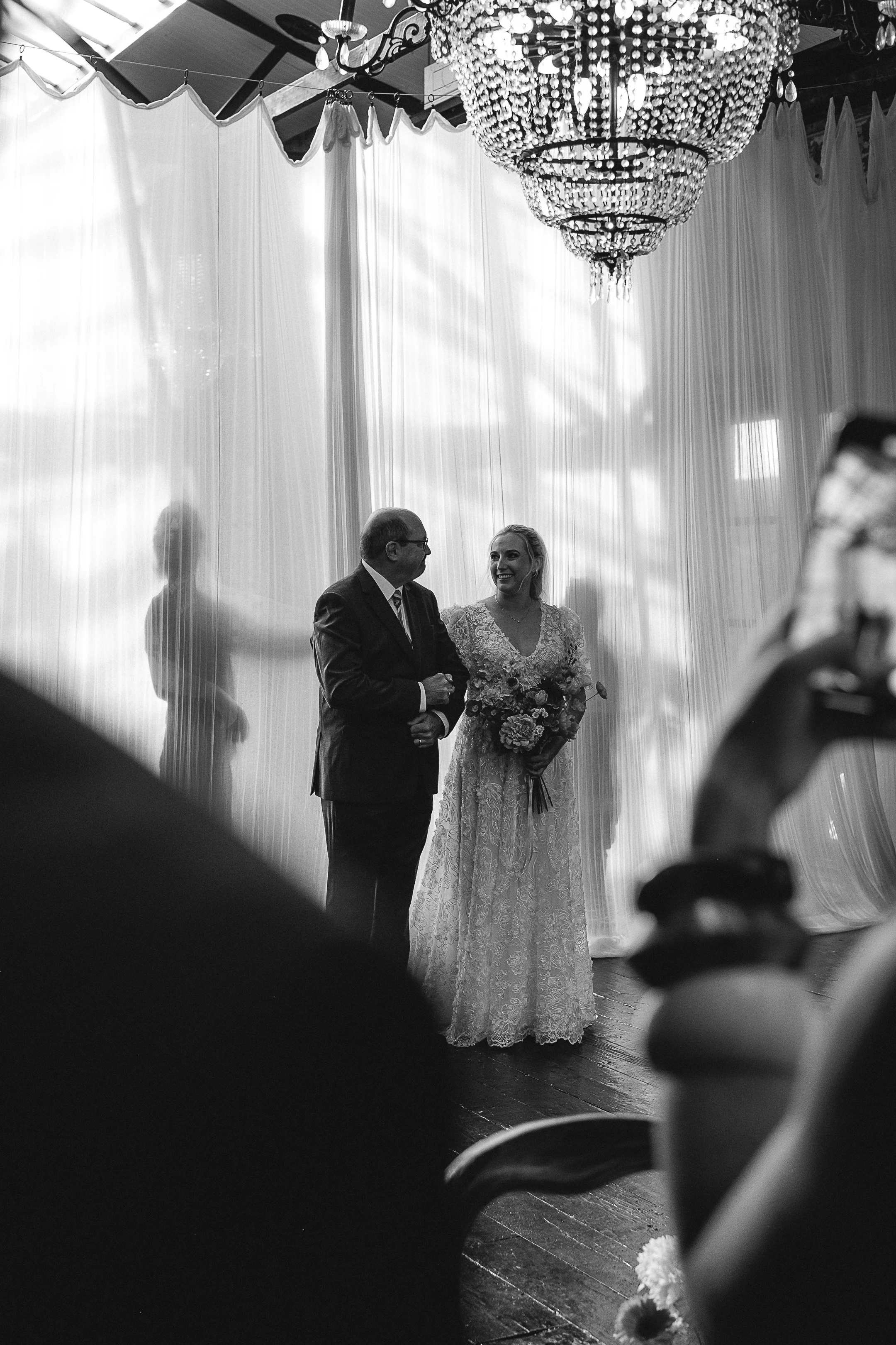 A bride in a lace wedding dress holding a bouquet, standing beside an older man in a suit, both smiling at each other during a wedding ceremony. Behind them is a sheer curtain with sunlight streaming through, and a large chandelier hanging from the c