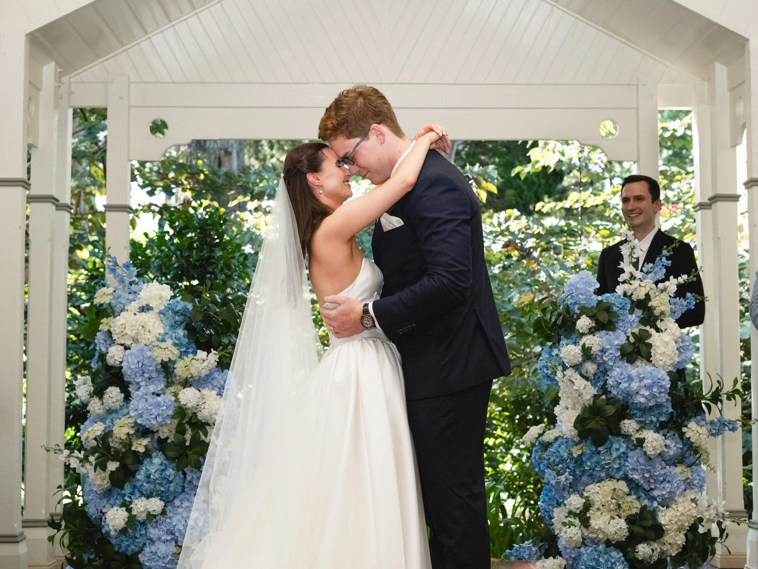 A bride and groom sharing a kiss during their wedding ceremony, standing under a white gazebo decorated with blue and white flowers, with a smiling best man in the background.