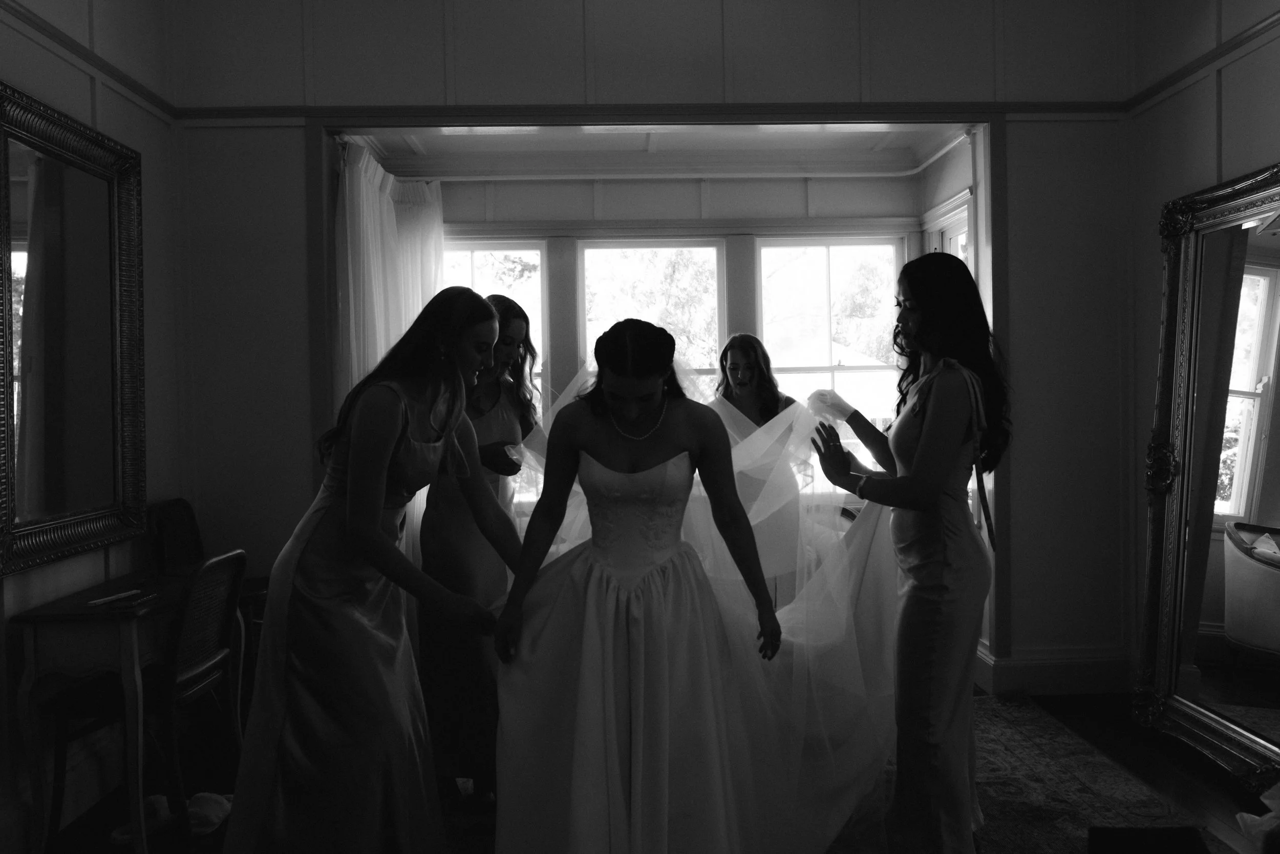 A bride and her bridesmaids praying together in a room with large windows and curtains, silhouetted by sunlight.
