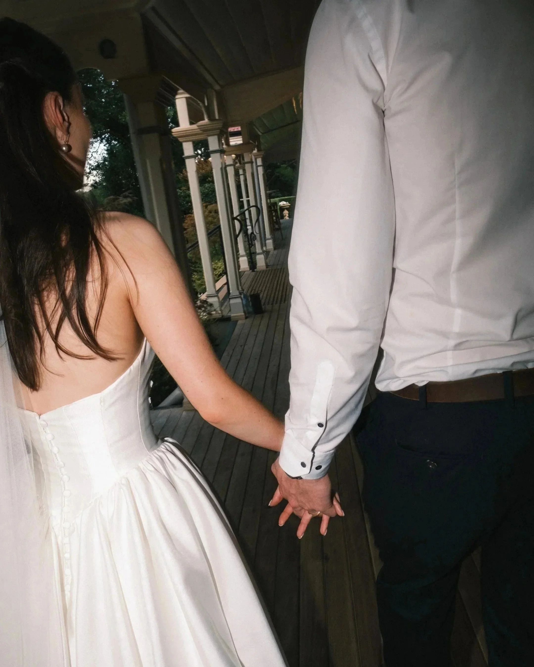 A bride and groom holding hands during their wedding, with the bride in a white wedding dress and the groom in a white shirt and dark pants, on a porch with a wooden floor and a house in the background.