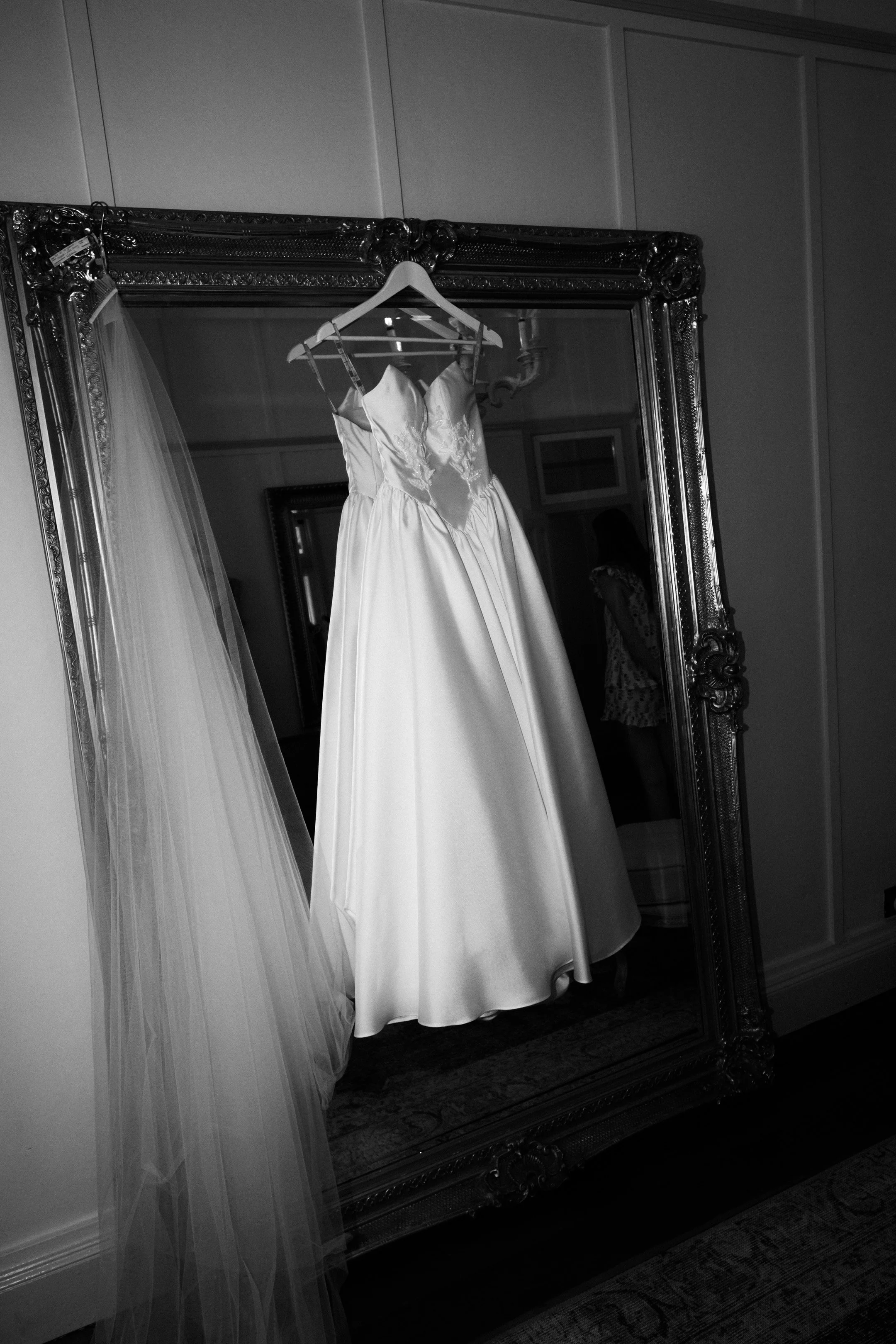A wedding dress hangs on a hanger in front of a large, ornate mirror, with a veil draped next to it.