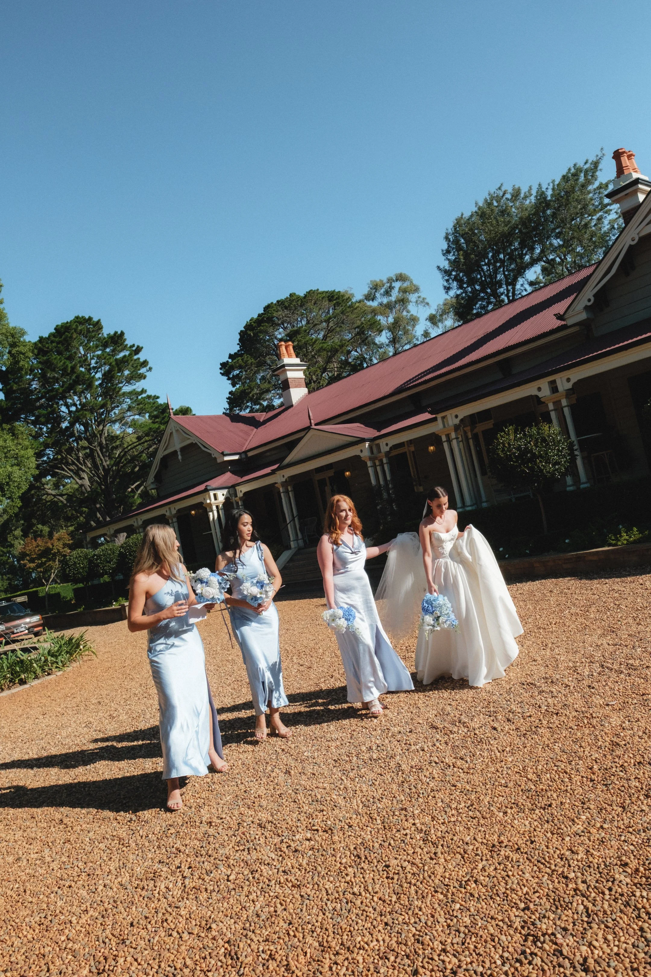 A bride and her bridal party, consisting of three bridesmaids, walking outside on a gravel path near a large house with a red roof on a sunny day.