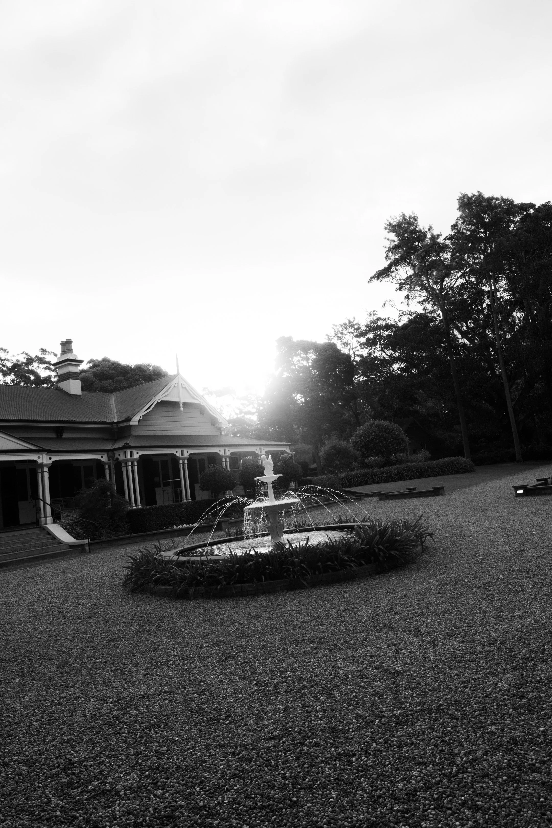 A black and white photo of a house with a fountain in front, surrounded by trees and a gravel lawn, with sunlight in the background.