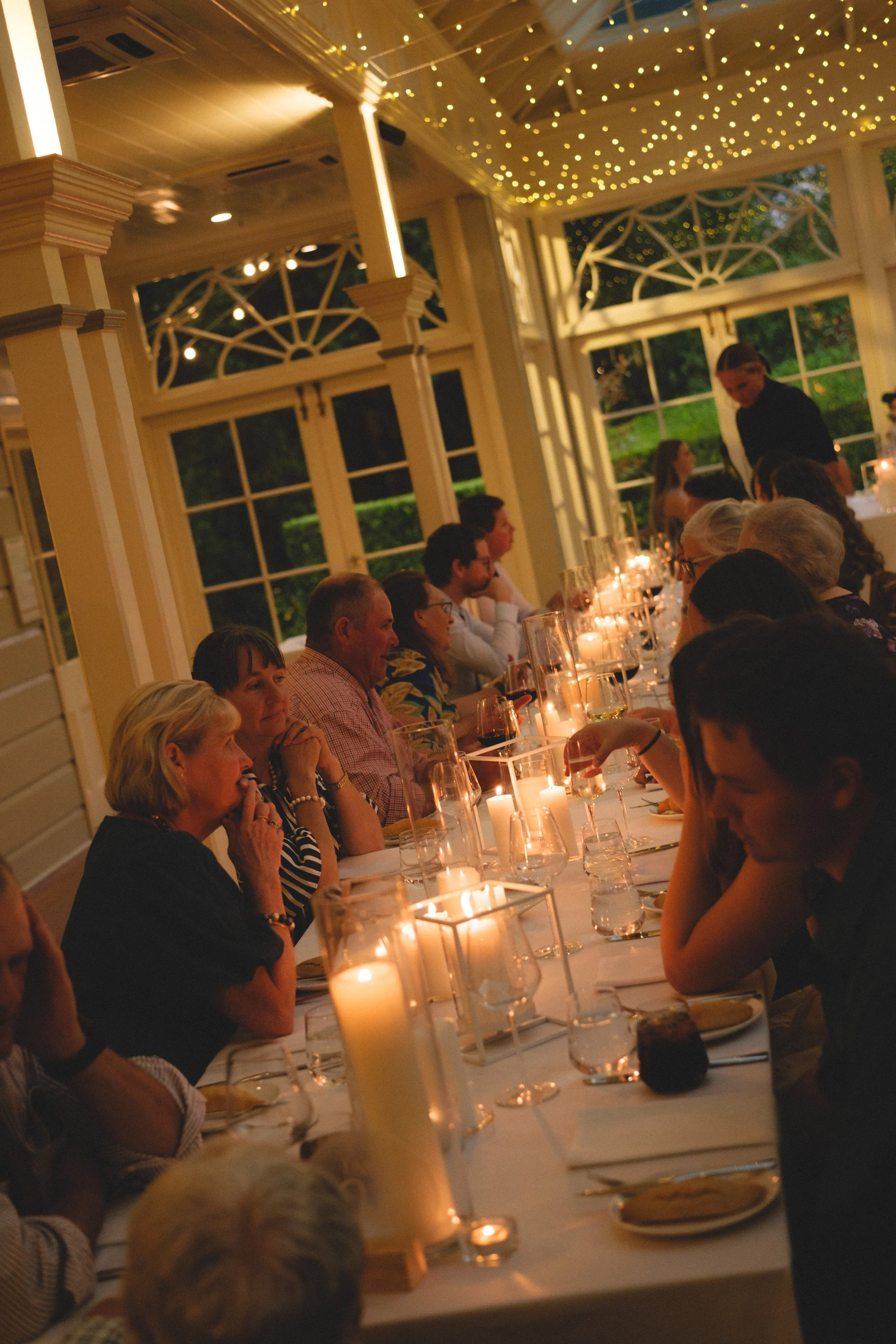 People seated at a long dinner table decorated with candles in a well-lit room with fairy lights on the ceiling and large windows showing the outside greenery.