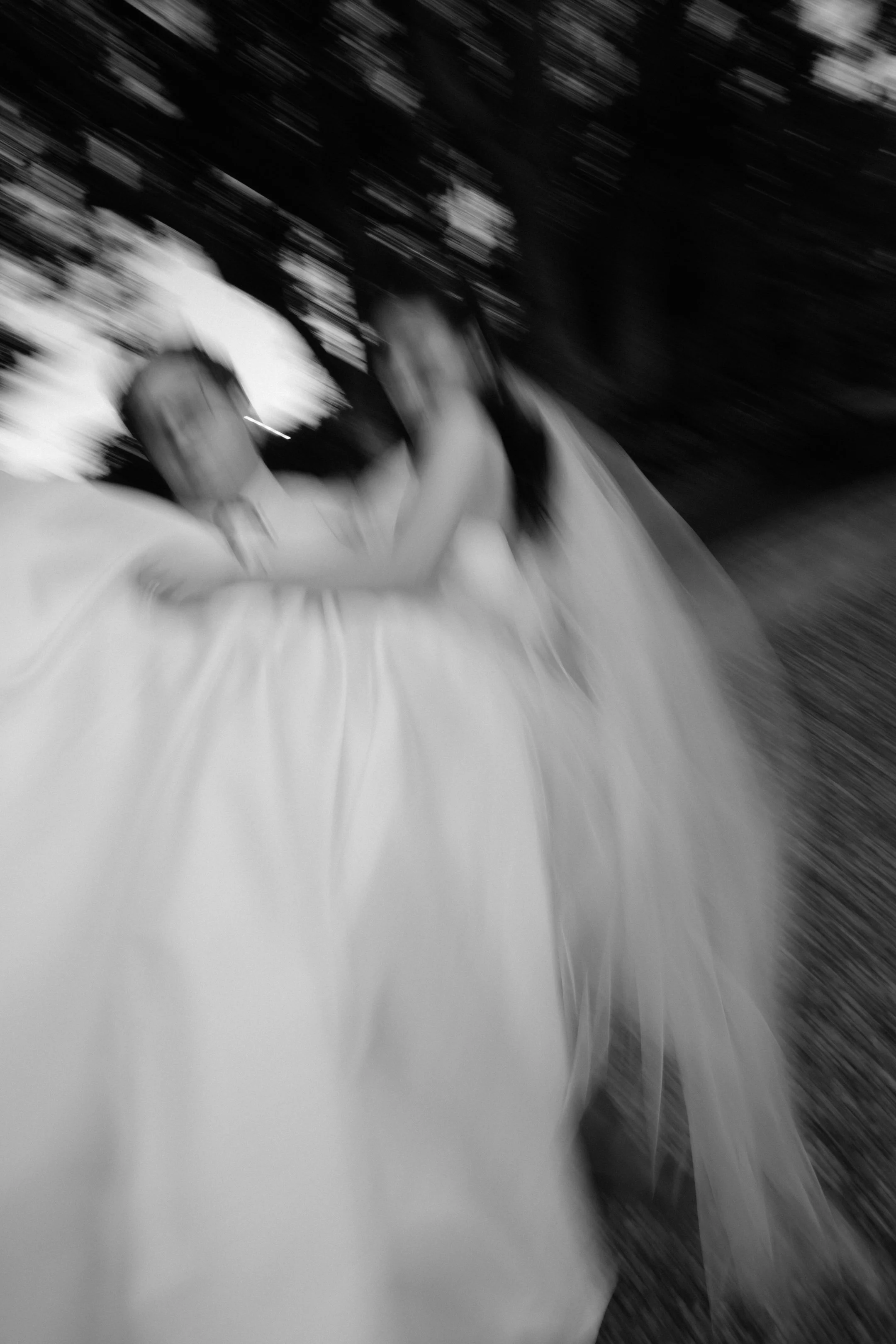 Black and white photo of a person in a wedding dress and a man, with a motion blur effect, outdoors with trees in the background.