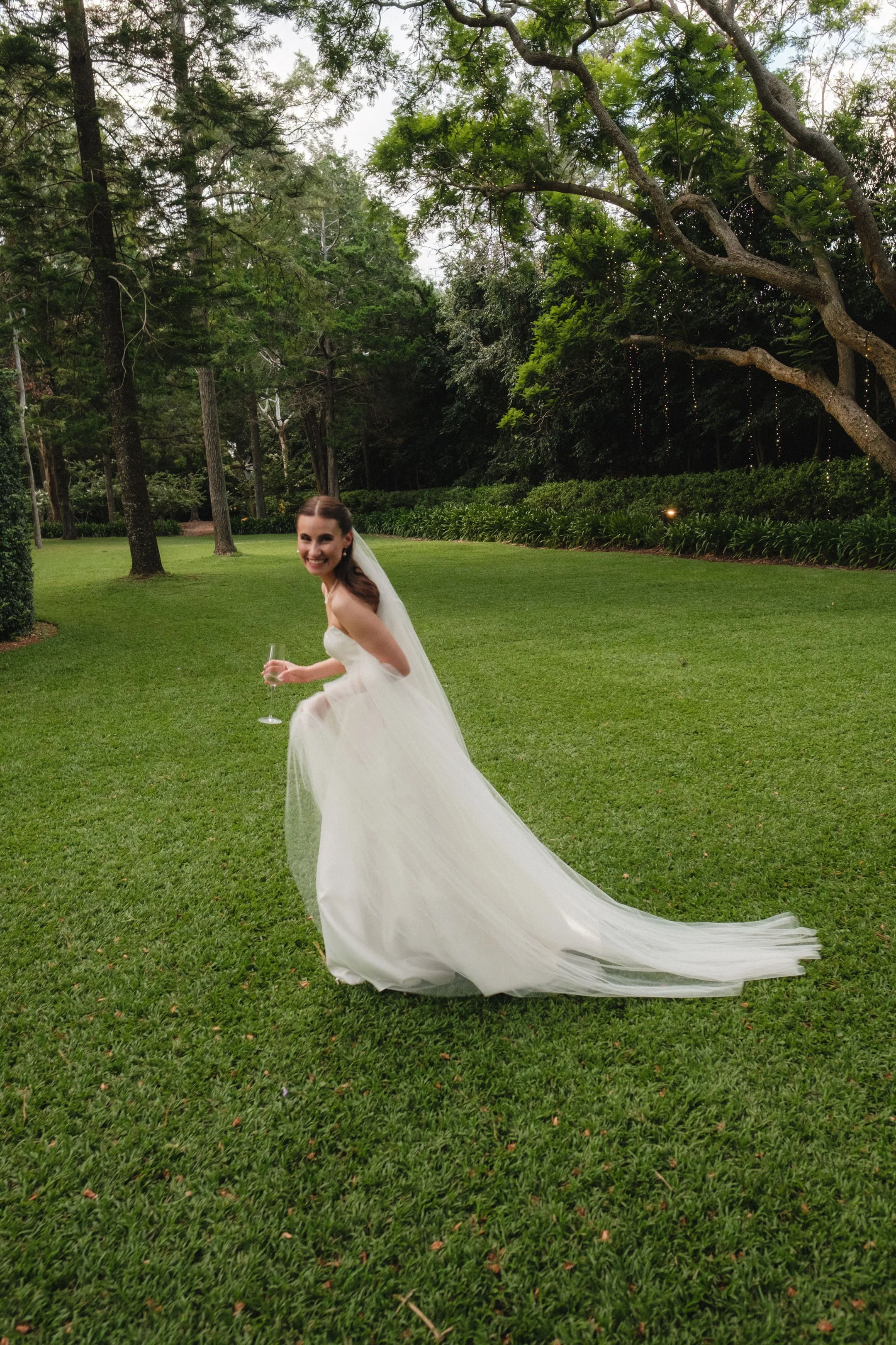 A smiling bride in a white wedding dress holding a champagne glass, standing on a lush green lawn with trees and bushes in the background.