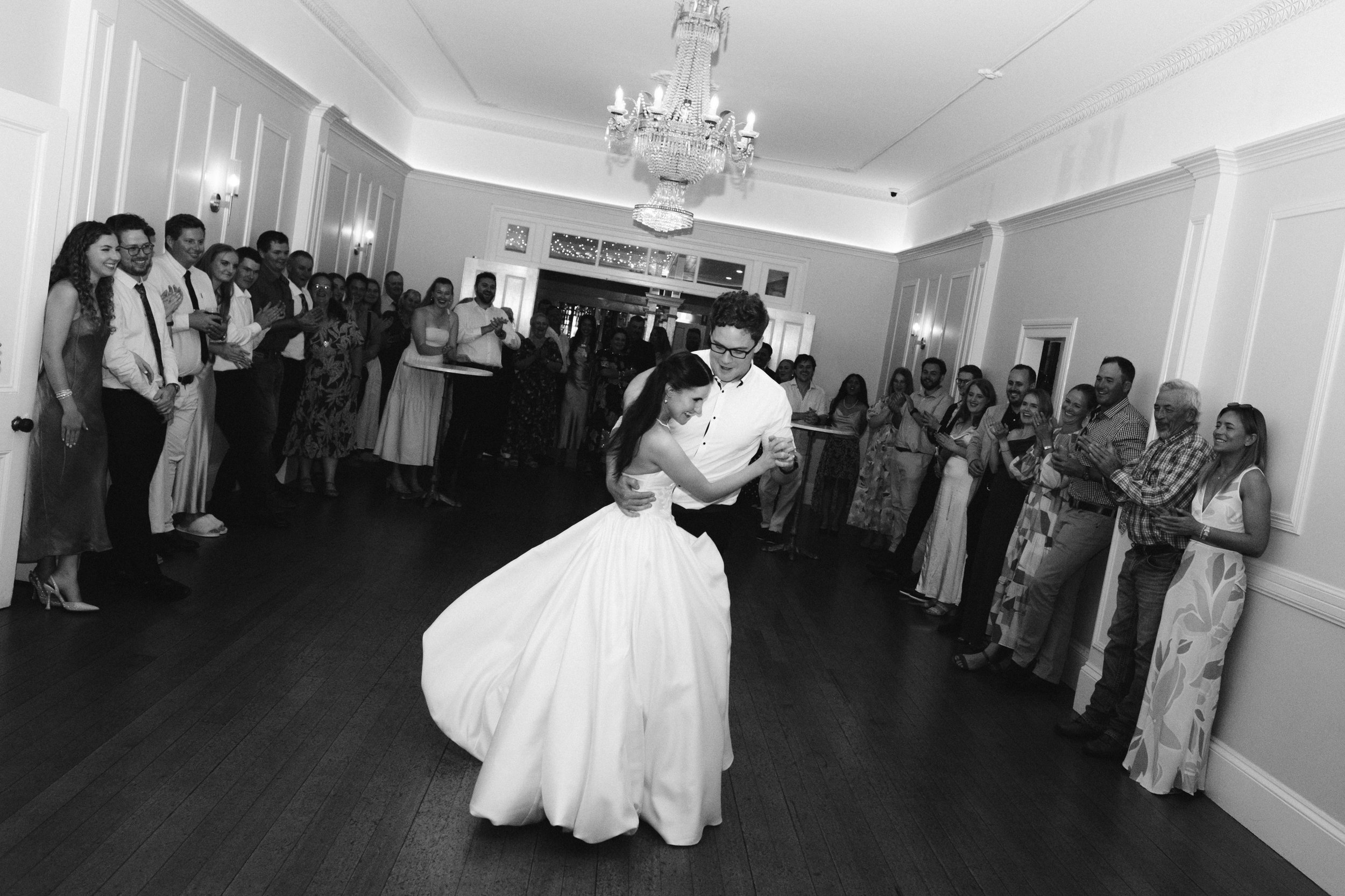 A couple is dancing in the center of a room with a large crowd of people watching and clapping around them. The woman is wearing a white wedding dress and the man is wearing a white shirt and dark pants. The room has elegant decor with a chandelier a
