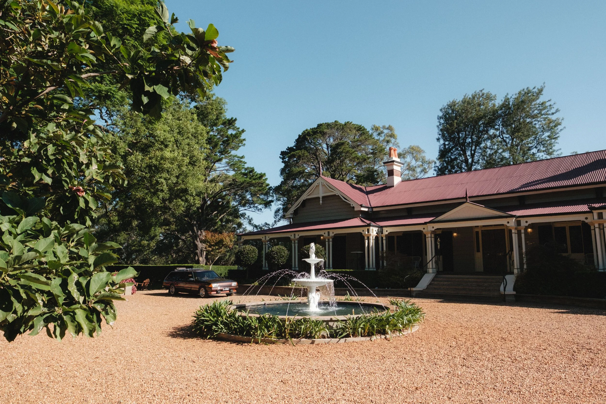 A large house with a red roof and porch, a fountain in the front yard, and a black station wagon parked on a gravel driveway, surrounded by trees and greenery under a clear blue sky.