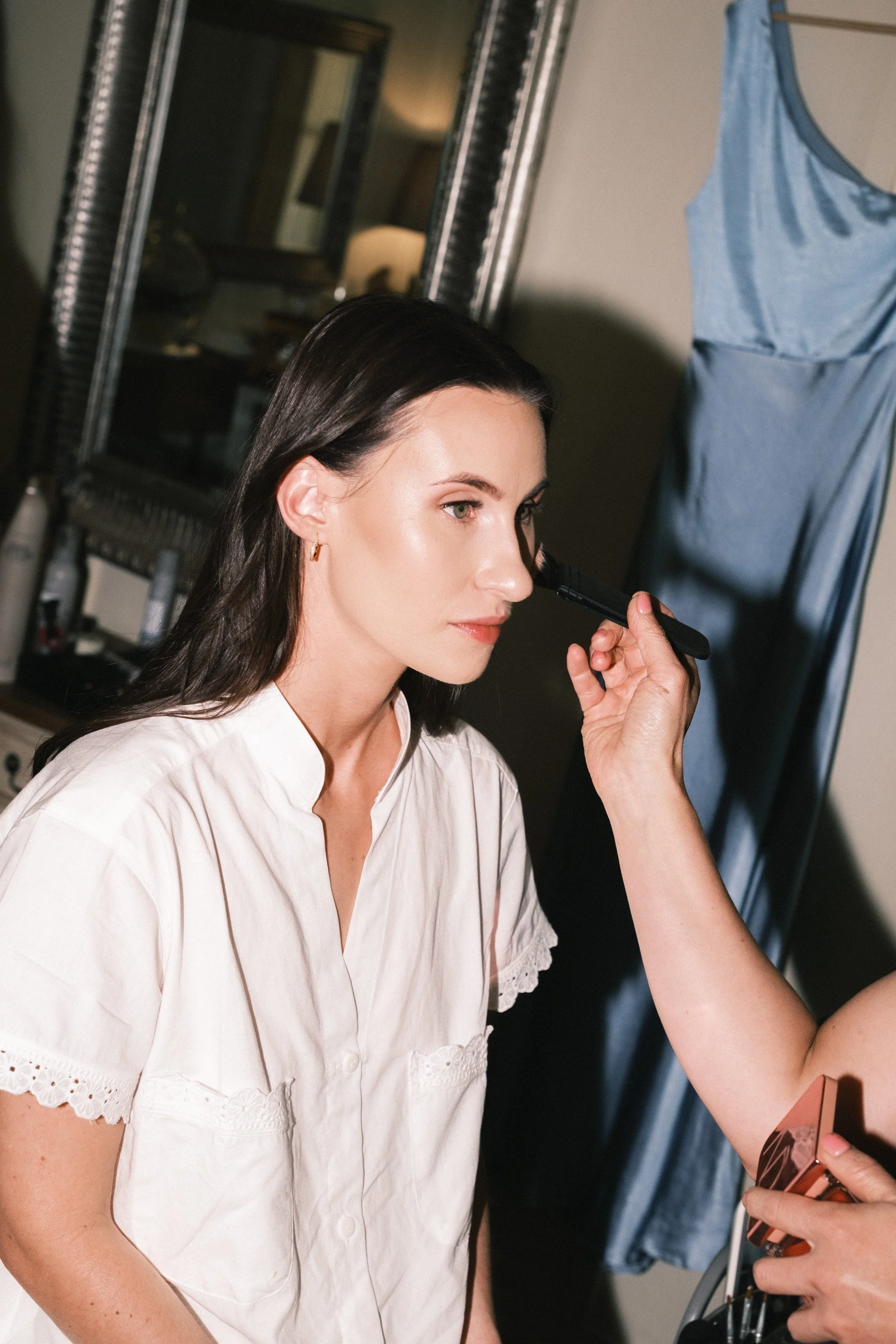 A woman with dark hair and light makeup has her face prepared as she is being makeuped by a person using a makeup brush, with a blue dress hanging on the wall behind her.