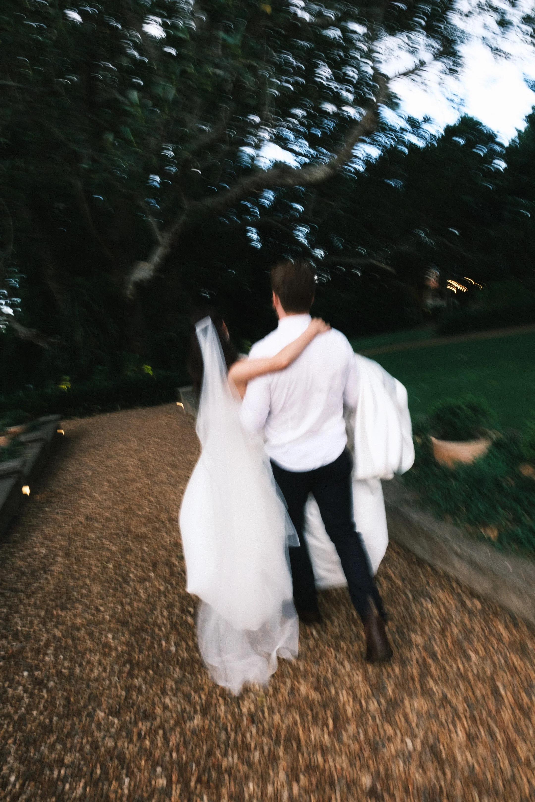 A bride and groom walking hand in hand on an outdoor pathway, surrounded by trees, seen from behind. The photo is blurred with motion, enhancing the romantic and dynamic feel.