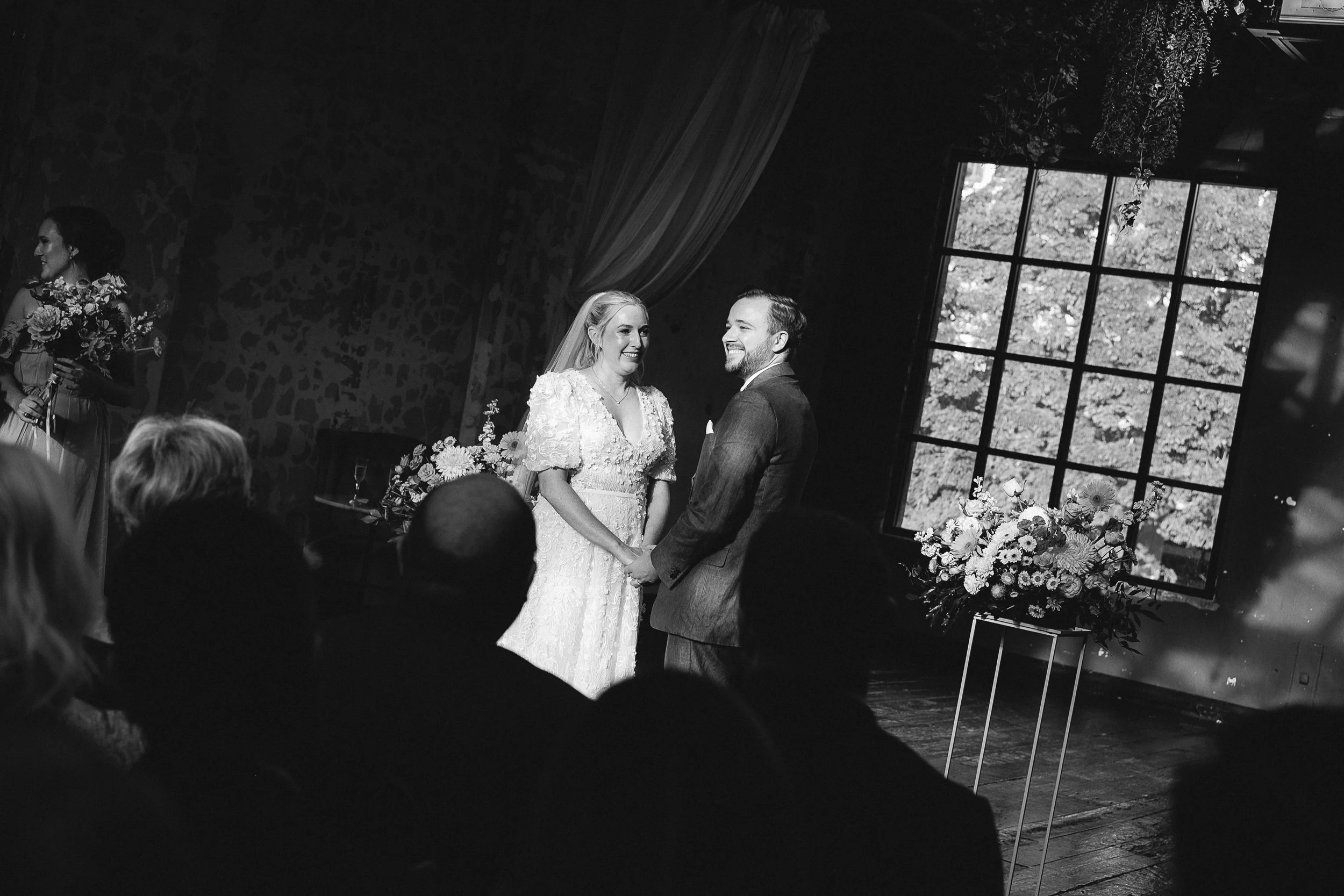 Black and white photo of a wedding ceremony, with a bride and groom holding hands and smiling at each other, in a room with a large window and floral decorations.