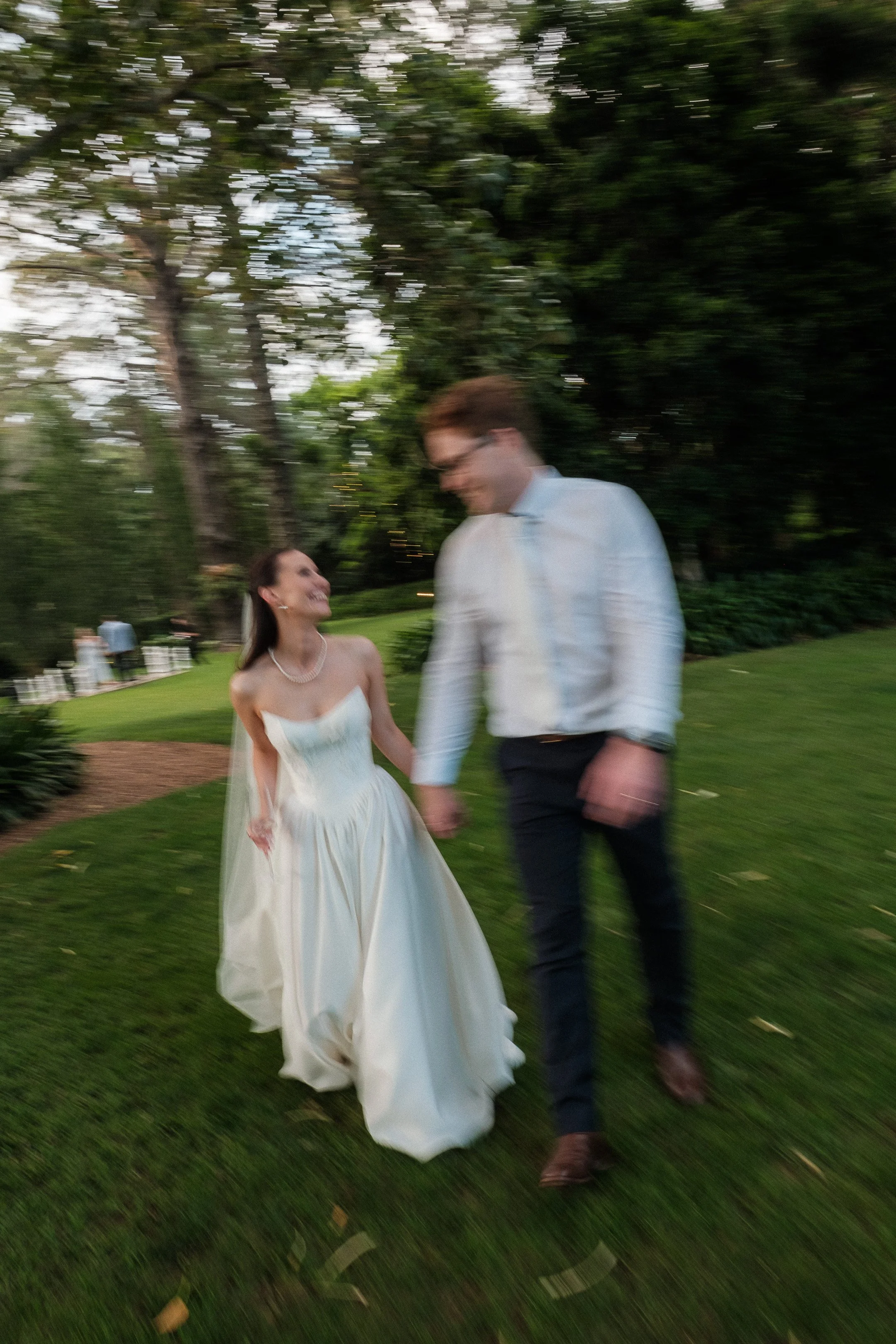 A bride and groom holding hands, walking and smiling in a lush green outdoor setting, possibly during their wedding celebration.