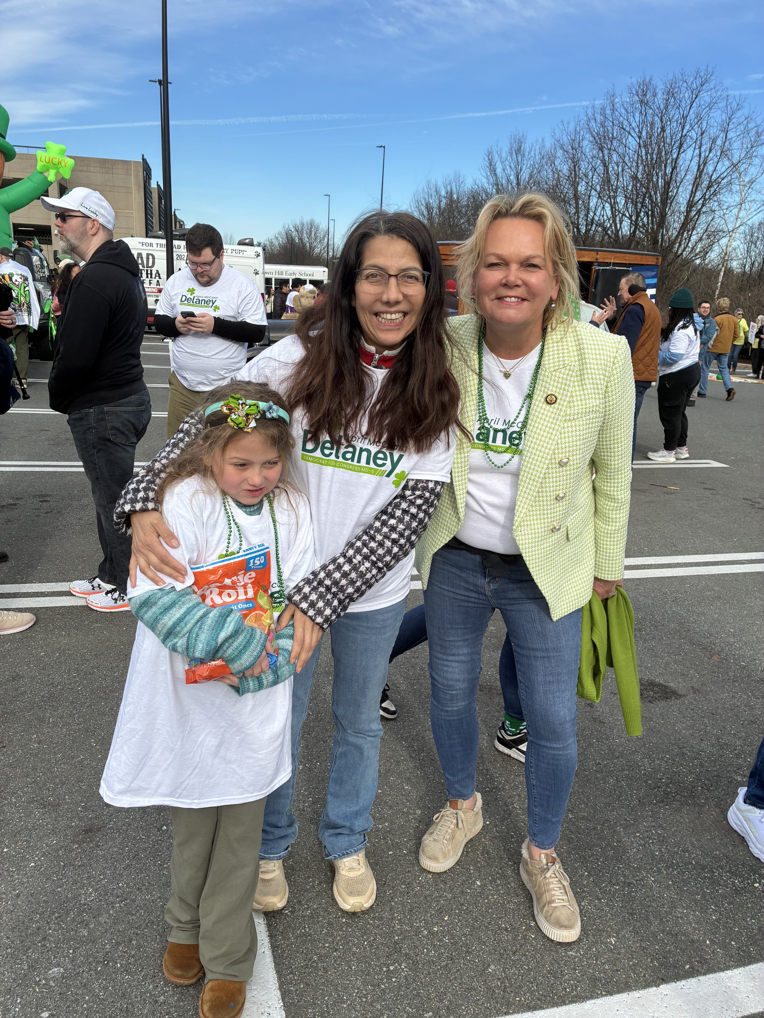 Three women and a girl smiling outdoors at a parade or event, wearing green-themed clothing and accessories.