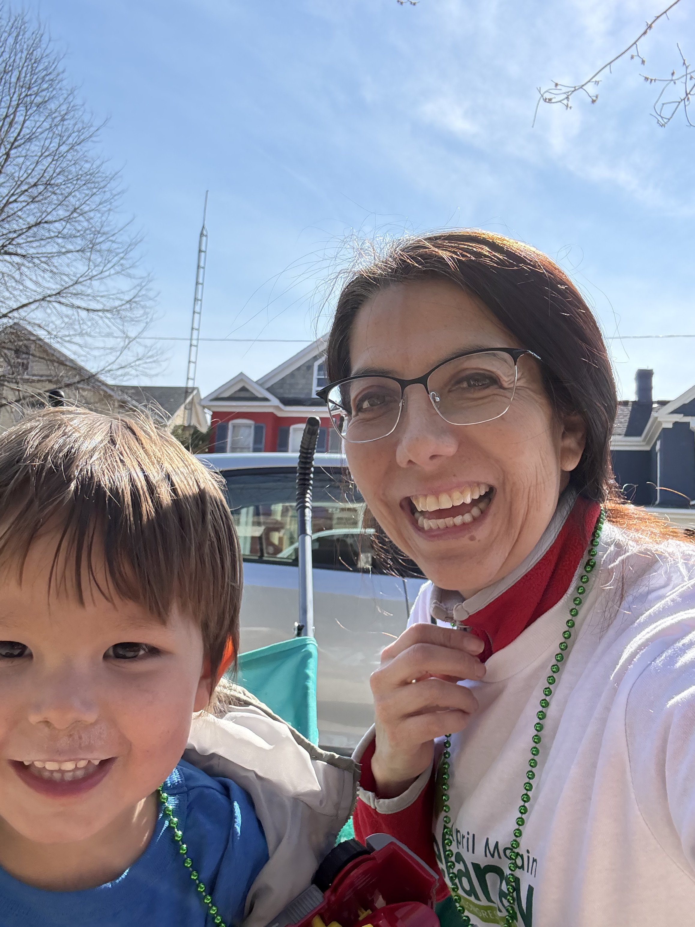 A smiling woman with glasses and a young boy are taking a selfie outside on a sunny day. The woman is wearing a white shirt, red scarf, and green bead necklace, while the boy is wearing a blue shirt and a jacket. They are standing in front of parked cars with houses in the background and clear blue sky above.