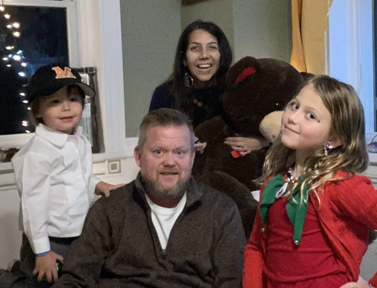 Family celebrating Christmas indoors, with a woman, a boy, a girl, and a man. The woman holds a large stuffed animal, and the children wear festive clothing. The scene has holiday decorations and a cozy atmosphere.