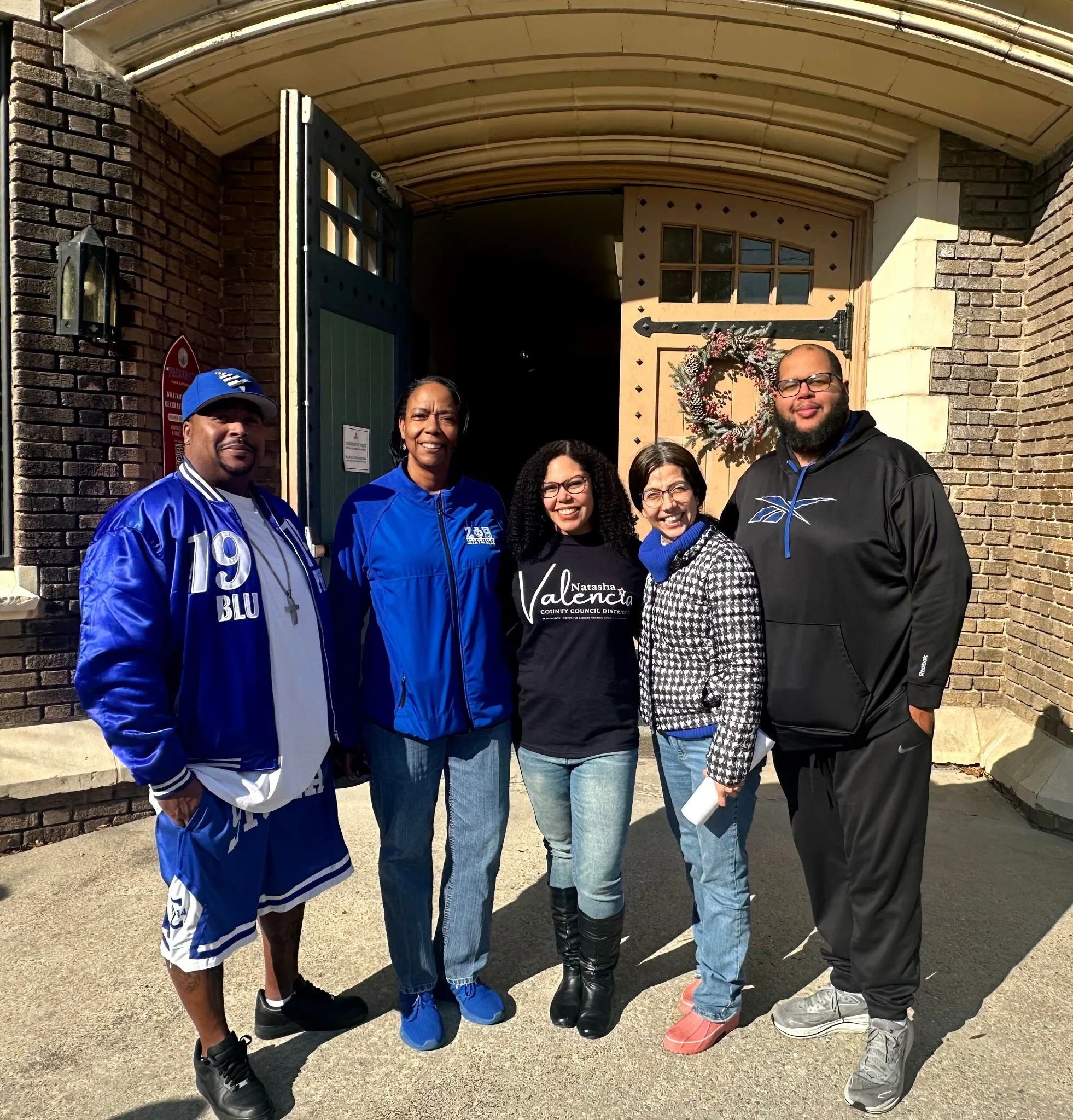 Group of five diverse people standing outside a brick building with a wooden door decorated with a holiday wreath, smiling at the camera.
