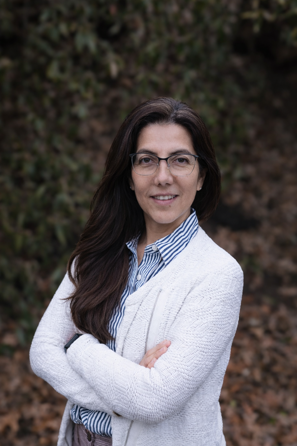 A woman with long dark hair, glasses, and a striped shirt under a white cardigan standing outdoors with a blurred natural background.