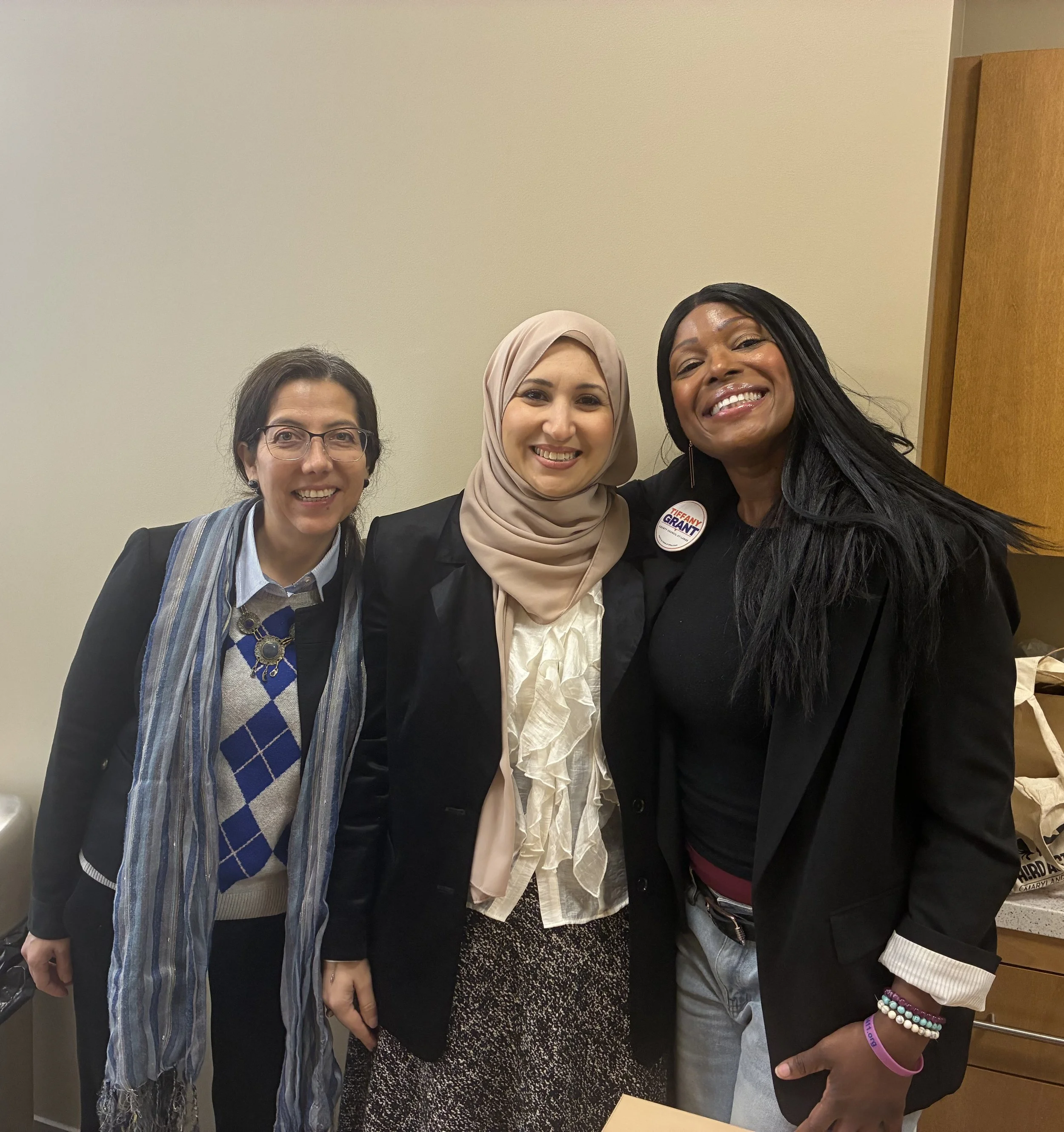 Three women standing together in an indoor setting, smiling at the camera. They are dressed in business casual attire, and one woman is wearing a button that says "Tiffany Grant."
