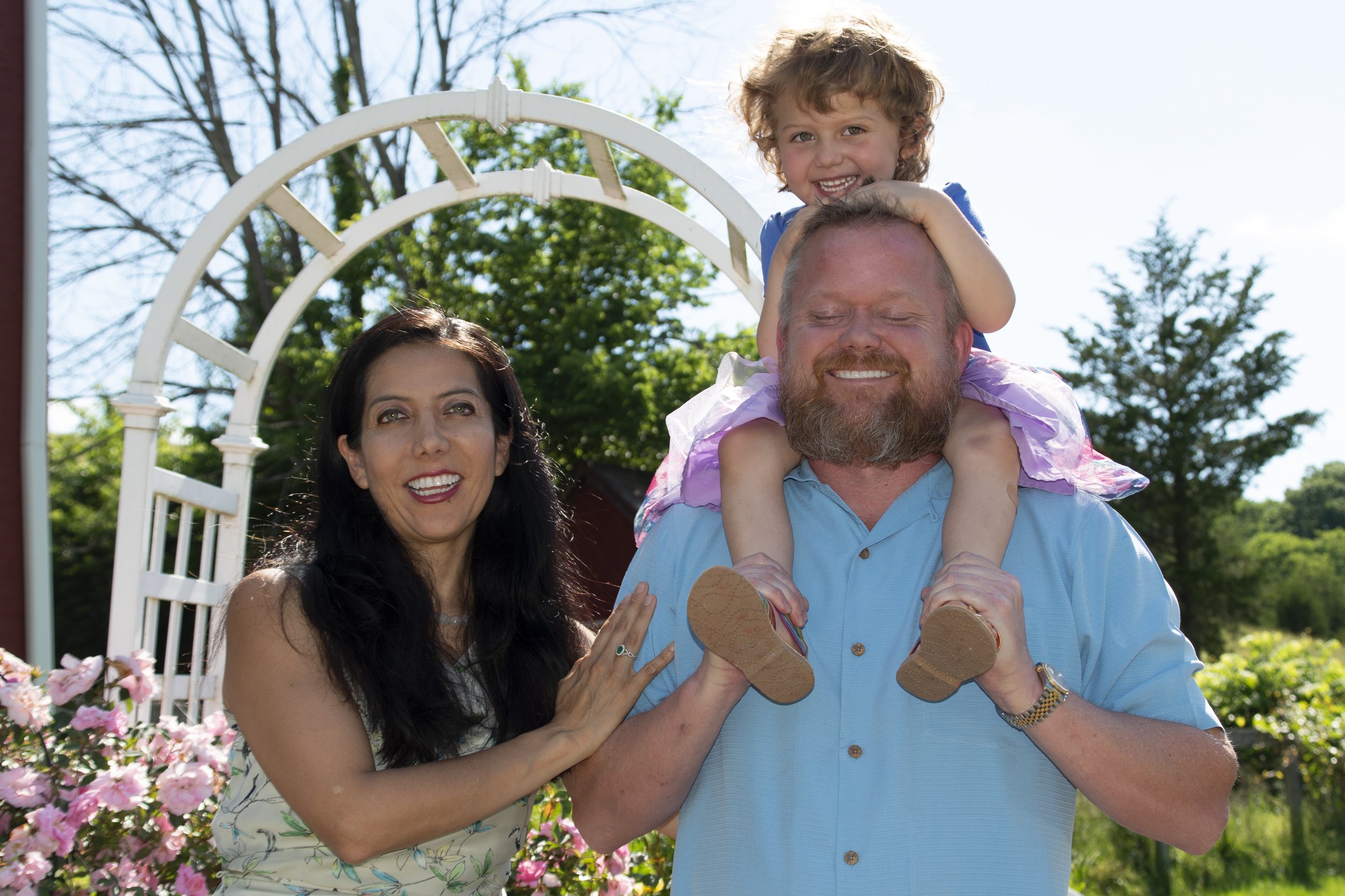 A happy family outdoors on a sunny day, with a woman with dark hair, a man with a beard, and a young girl with curly hair playing on the man's shoulders near pink flowers and greenery.