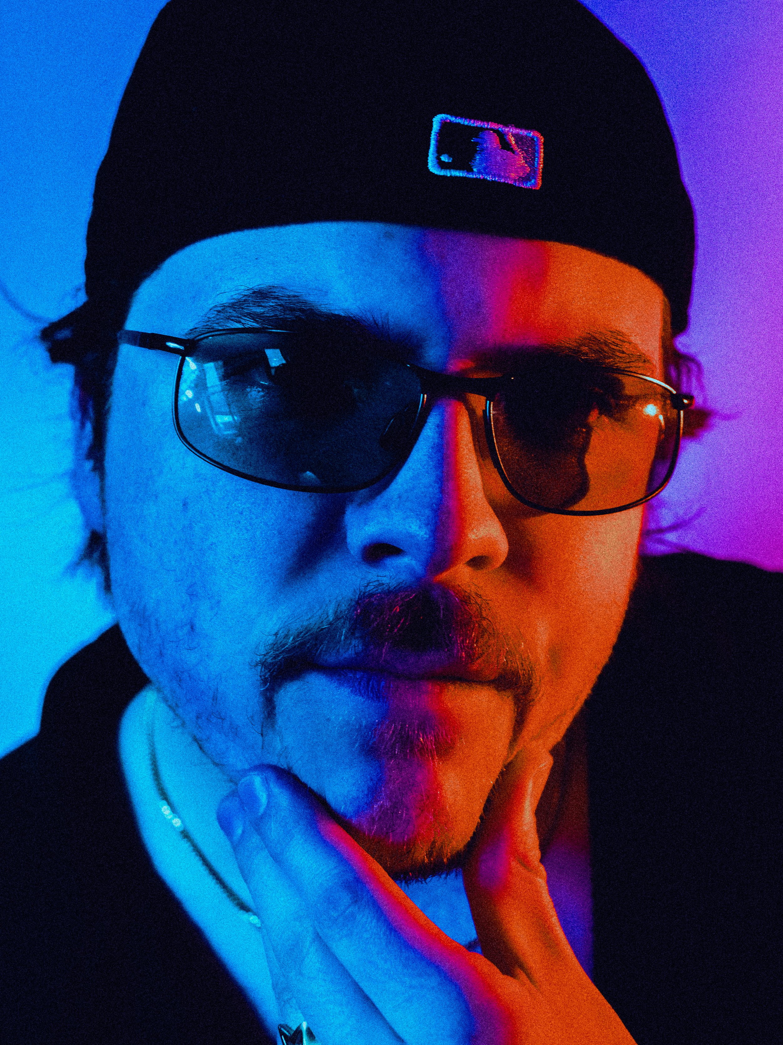 A person wearing a black beanie with a colorful logo, sunglasses, and a black hoodie, illuminated with blue and purple lighting.
