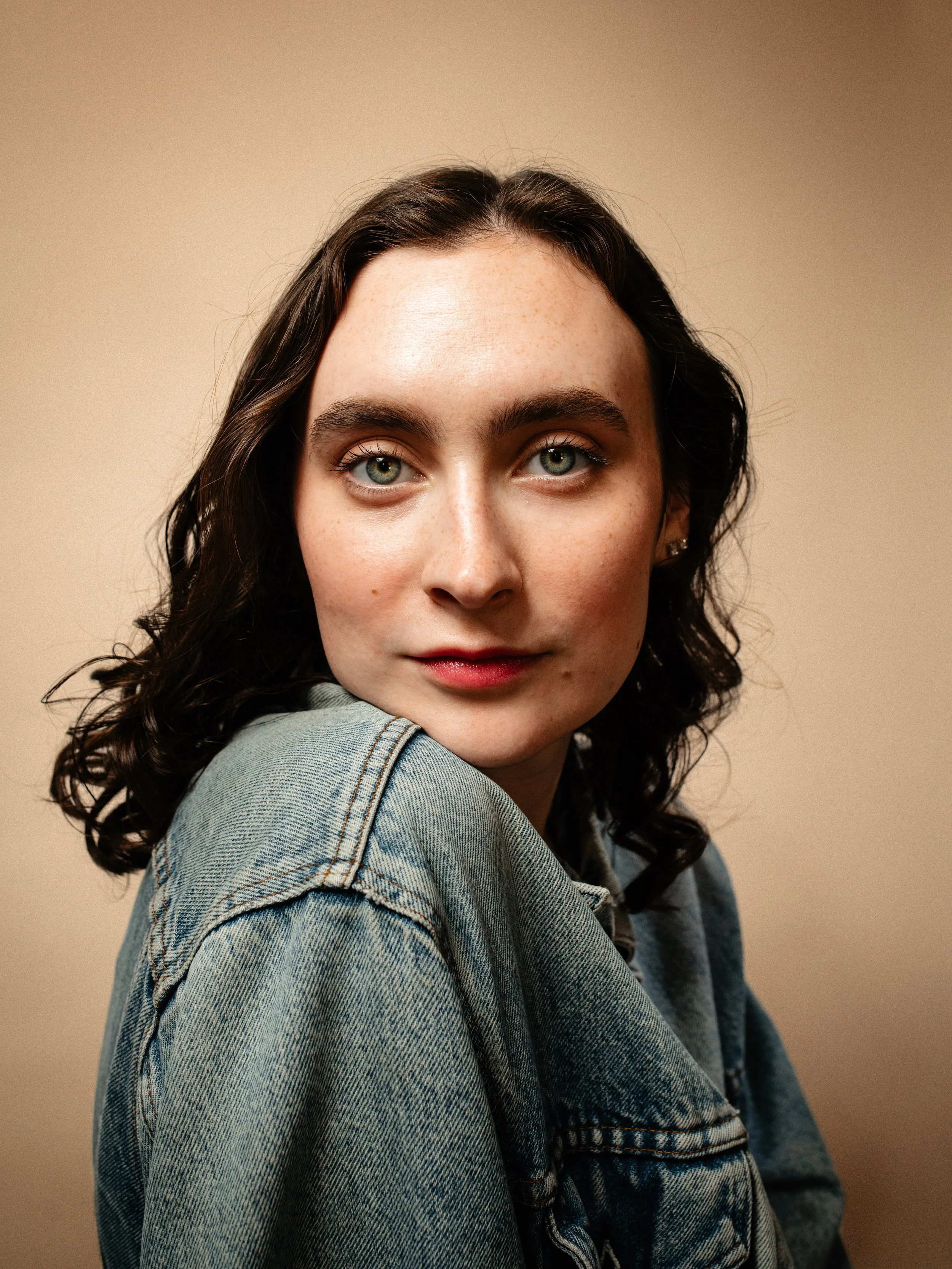 Close-up portrait of a young woman with dark wavy hair, blue eyes, wearing a denim jacket, looking at the camera against a neutral background.