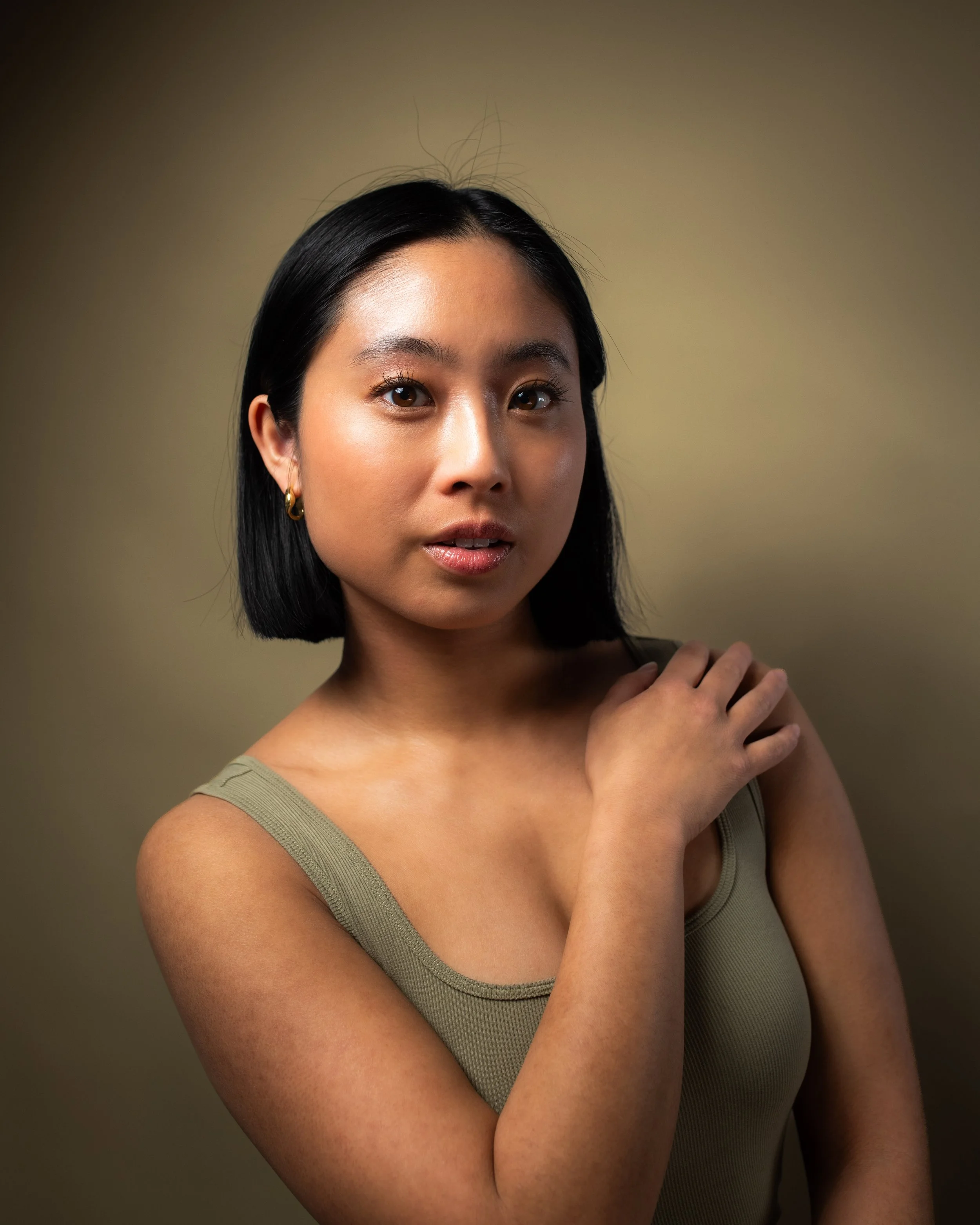 A young woman with medium skin tone, black shoulder-length hair, and gold hoop earrings looking directly at the camera with a neutral expression. She is wearing a sleeveless olive-green top and has her left hand resting on her right shoulder against 