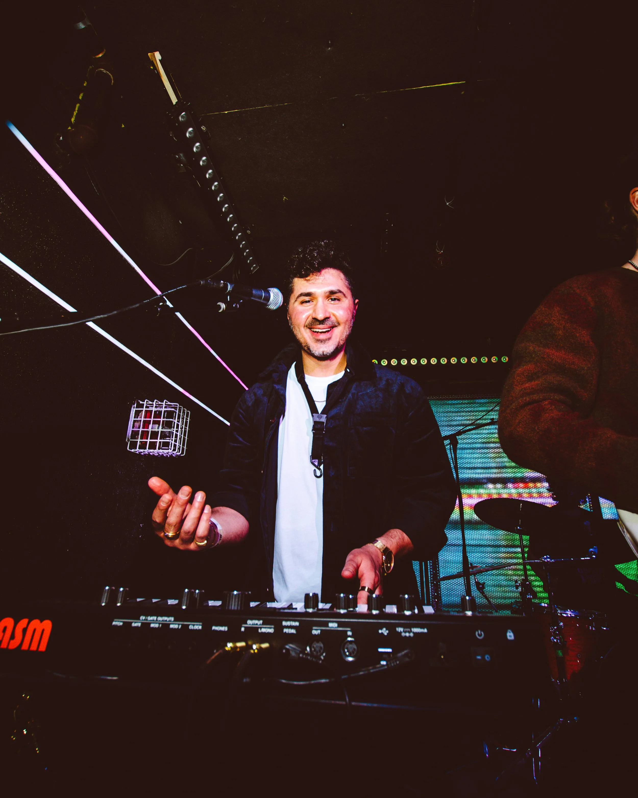 A DJ is smiling and playing music at a nightclub, standing behind a DJ mixer, with colorful neon lights in the background.