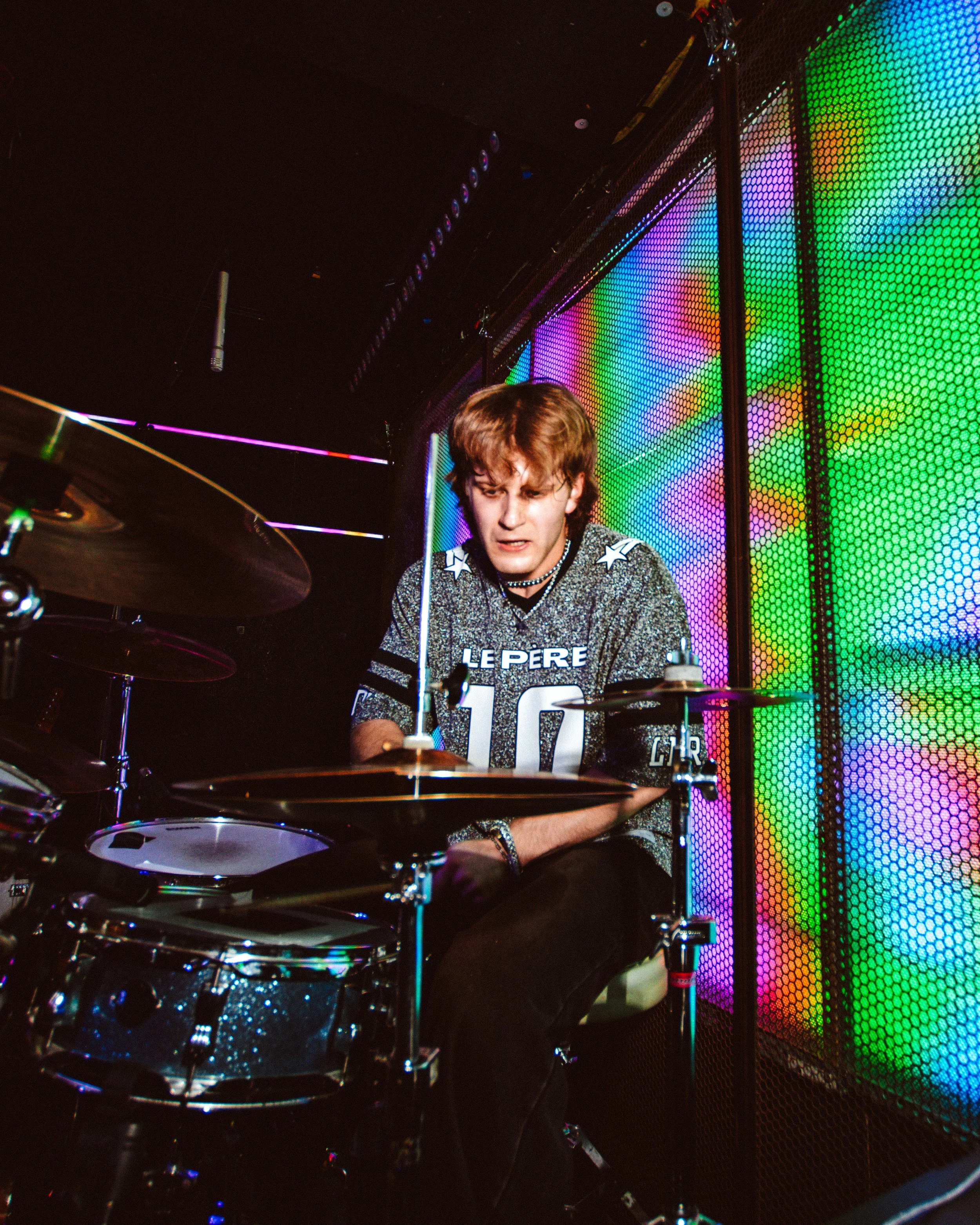 A young male drummer playing in a nightclub with brightly colored LED walls behind him, wearing a metallic shirt with the number 10 on it.