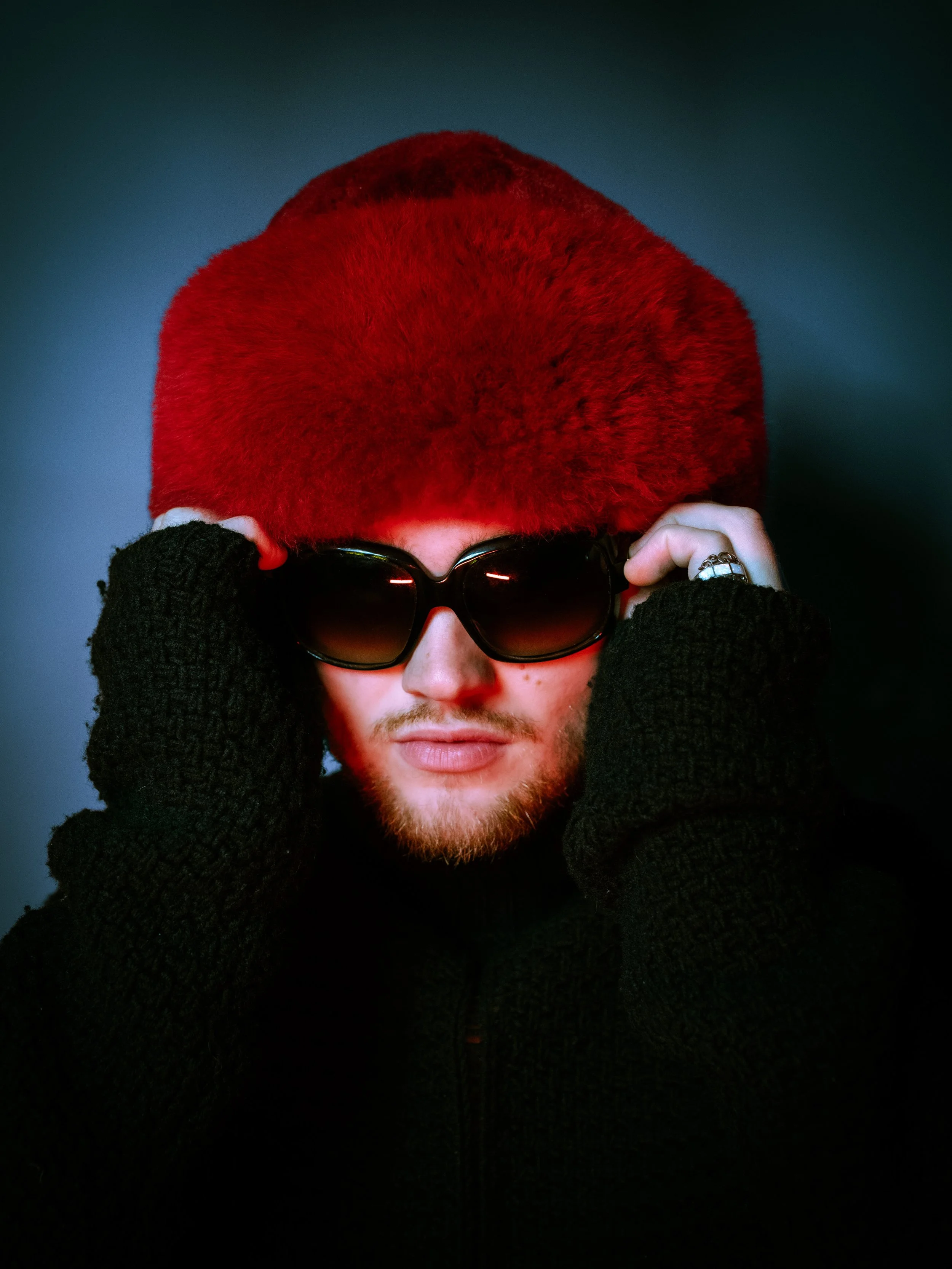 Person wearing large sunglasses, a fuzzy red hat, black gloves, and a black outfit, adjusting their glasses with both hands against a dark background.
