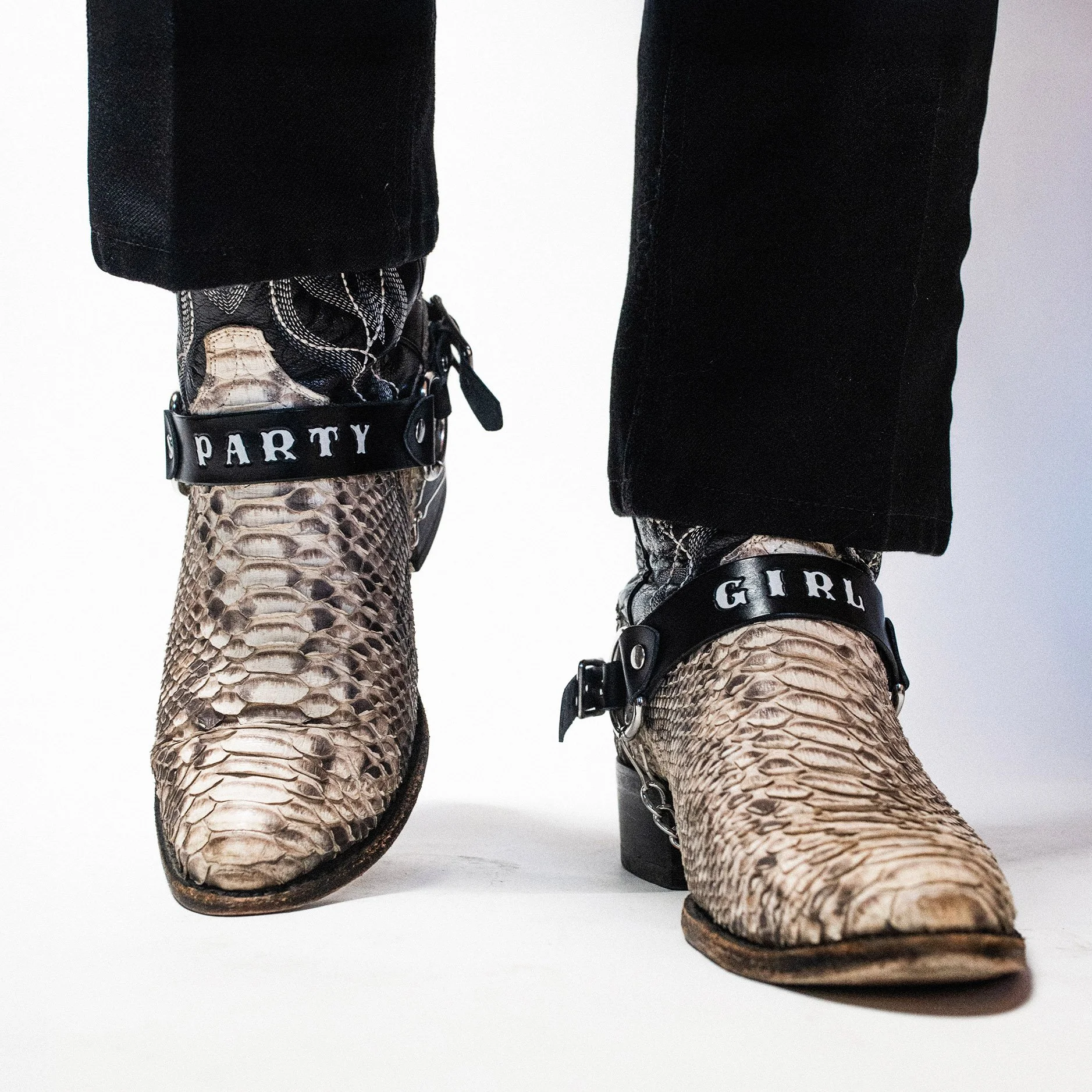 Person wearing snakeskin cowboy boots with decorative straps that say 'PARTY' and 'GIRL', black pants, and patterned socks.