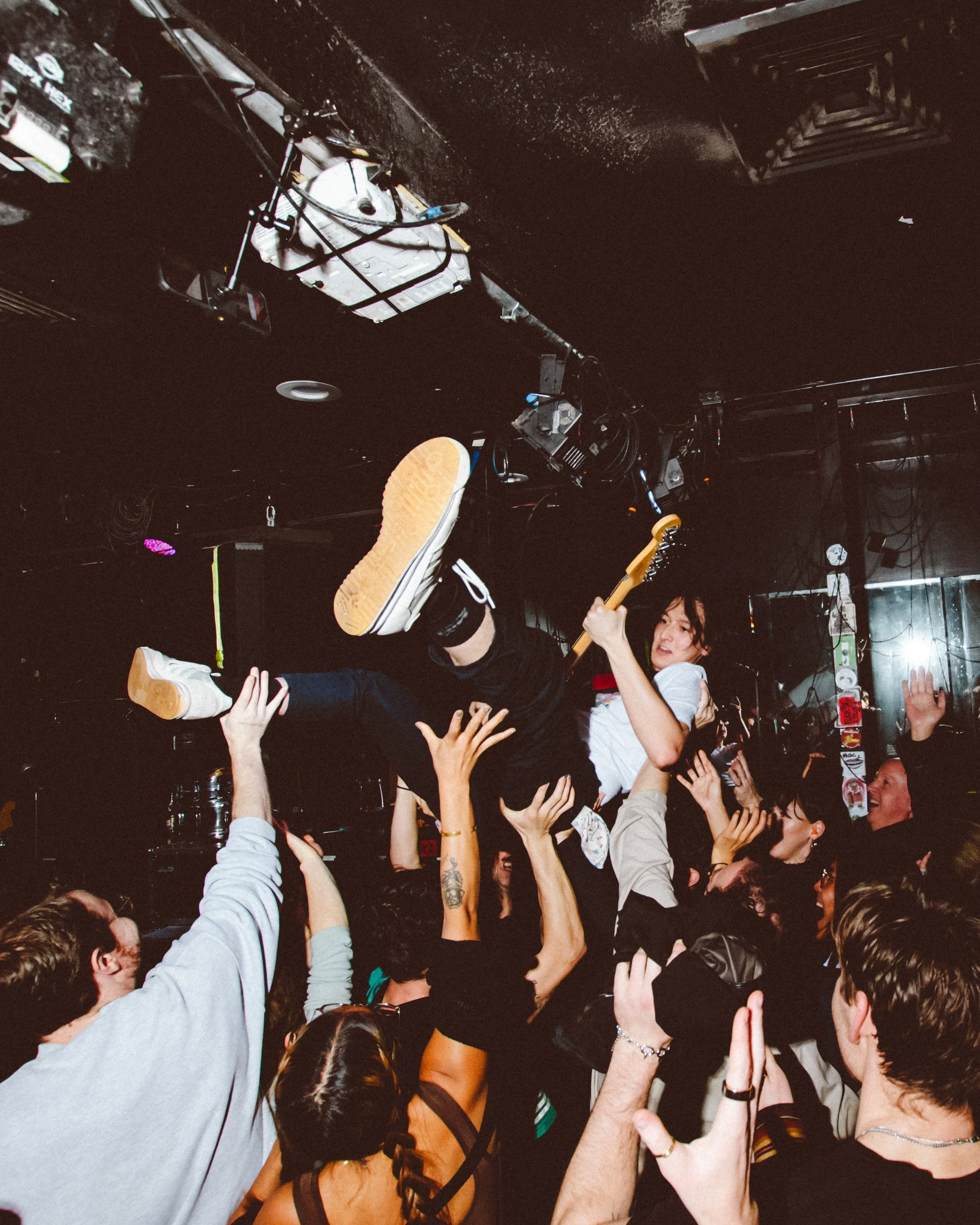 A woman crowd surfing at a concert, held up by several people's hands, with a guitarist playing on stage in the background.