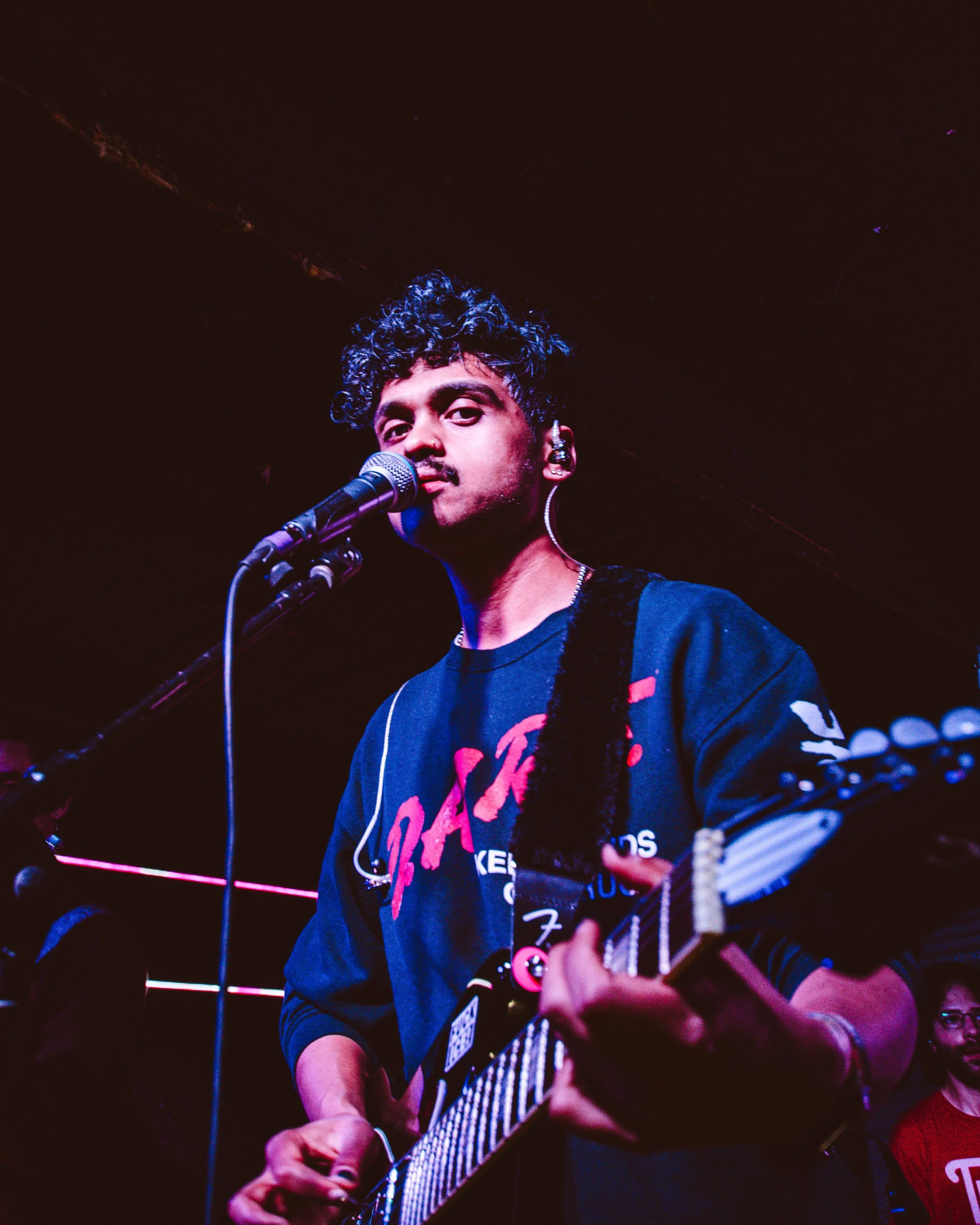 A musician performing on stage with a microphone and guitar, wearing a black sweatshirt with pink and white lettering, in a dark venue with pink lighting.
