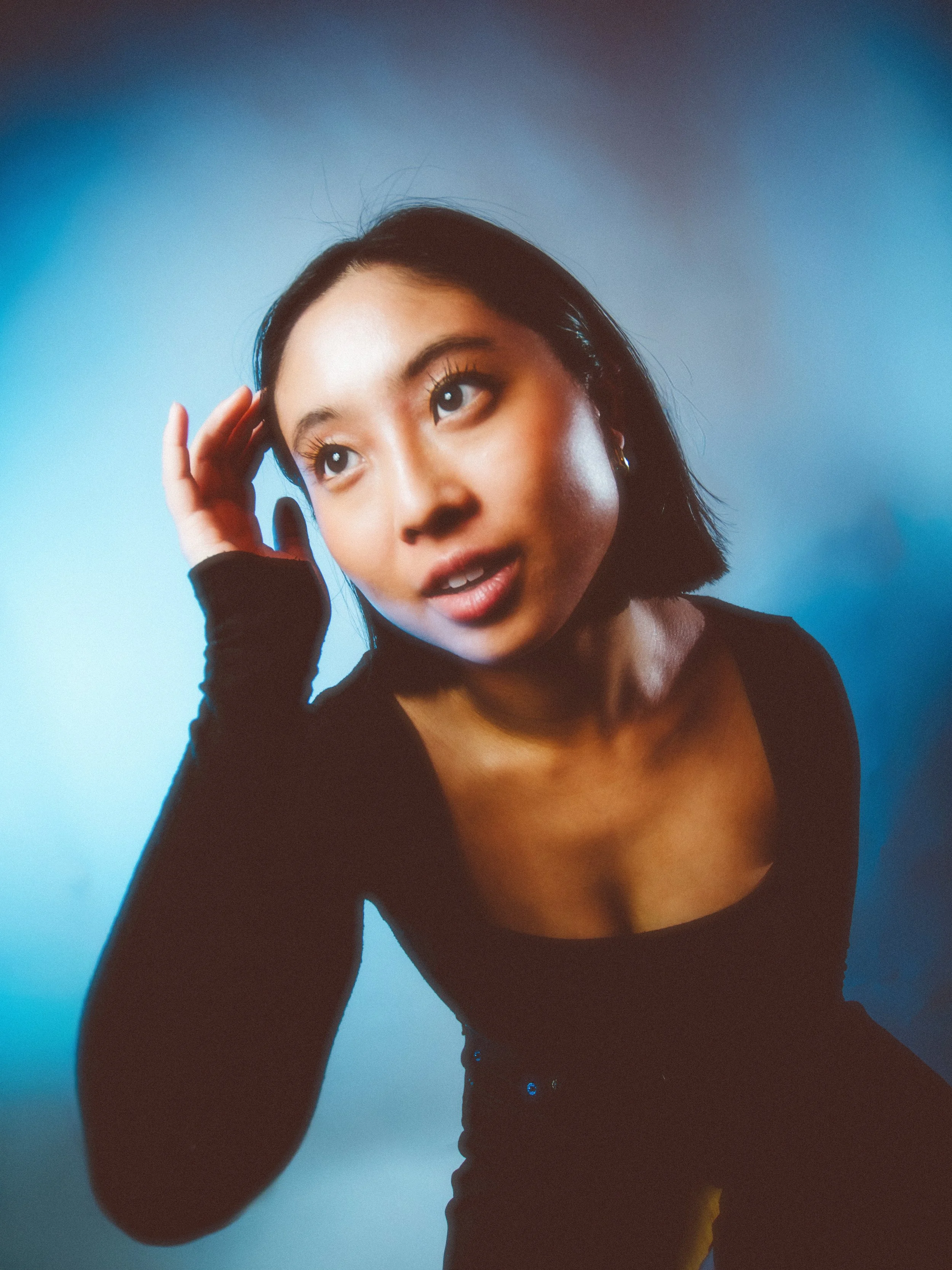 A young woman with short black hair and light skin, wearing a black top, touching her hair and looking up with a curious expression against a blue gradient background.