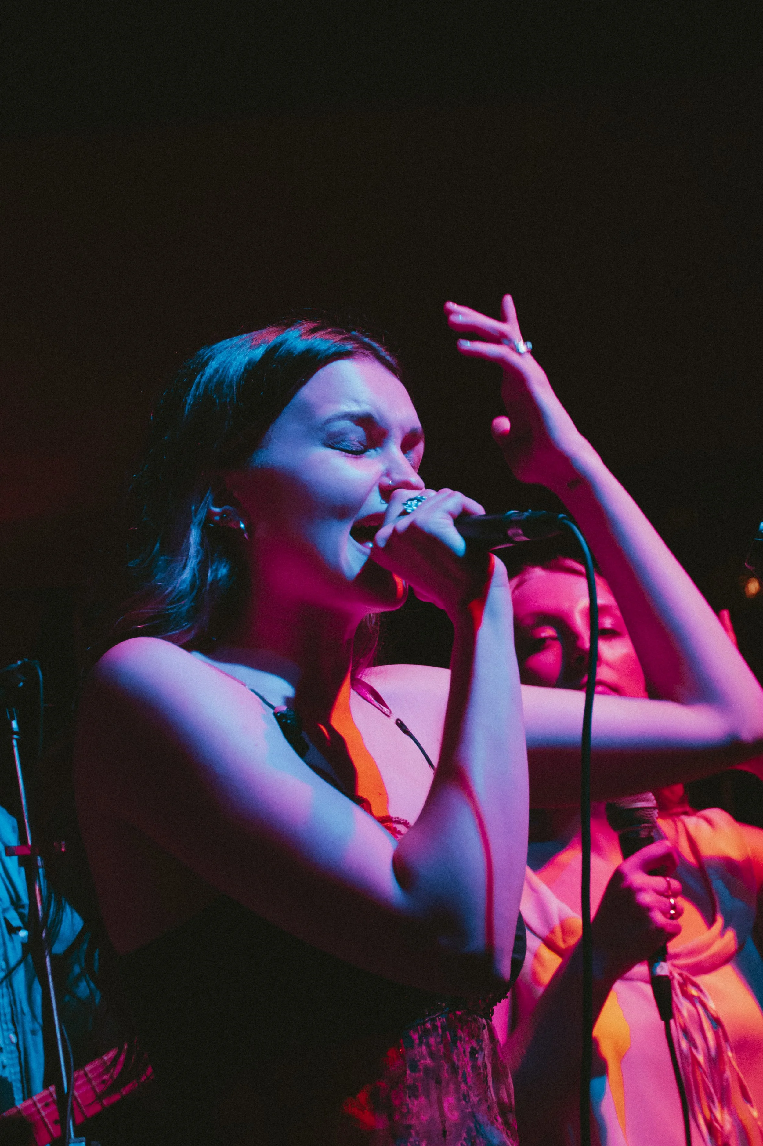A woman singing into a microphone on stage with her eyes closed and hand raised, illuminated by colorful stage lights, with another woman in the background holding a microphone.