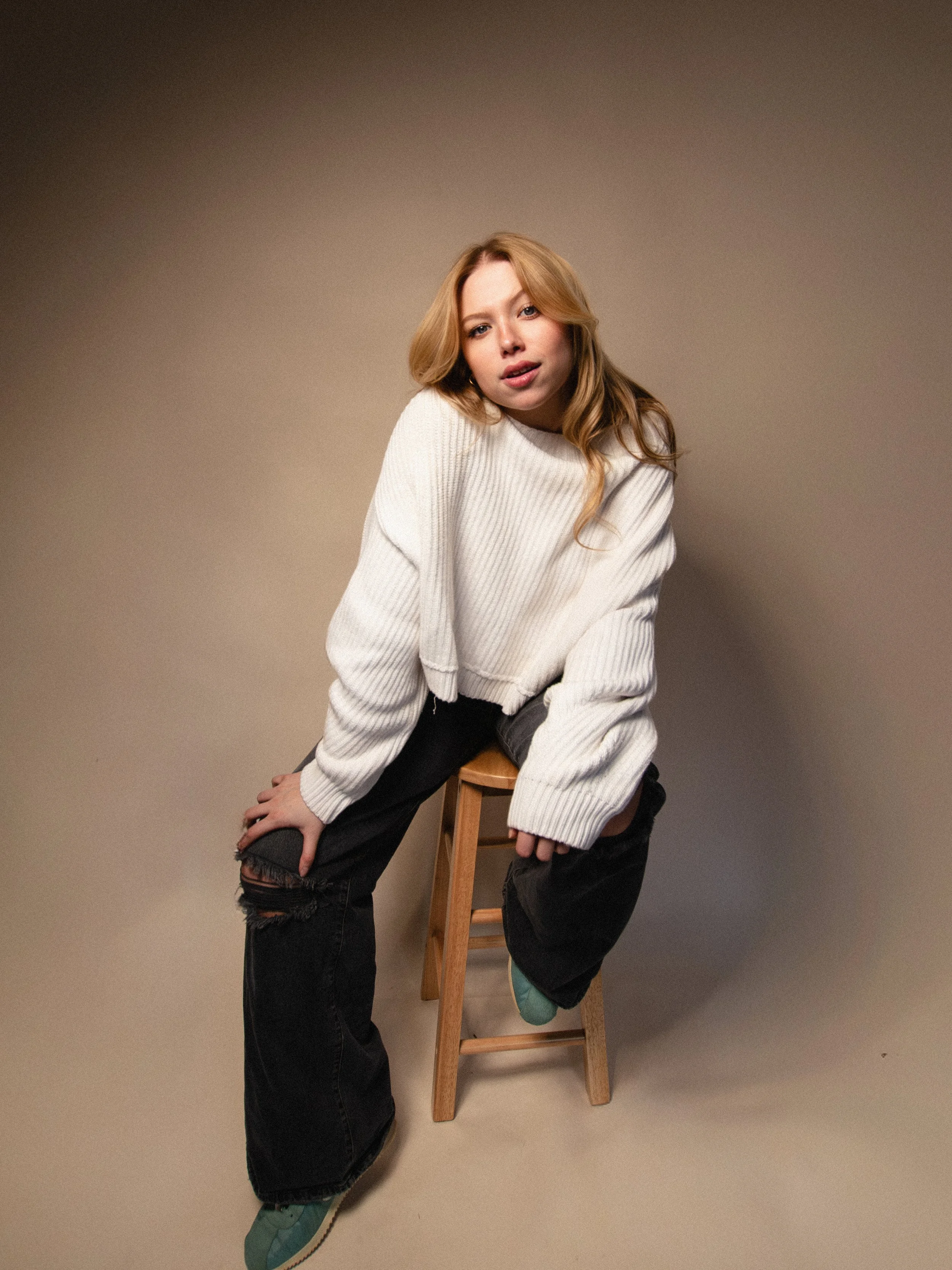 A young woman with long reddish-blonde hair sitting on a wooden stool against a plain background, wearing a white oversized sweater, black ripped jeans, and teal sneakers.
