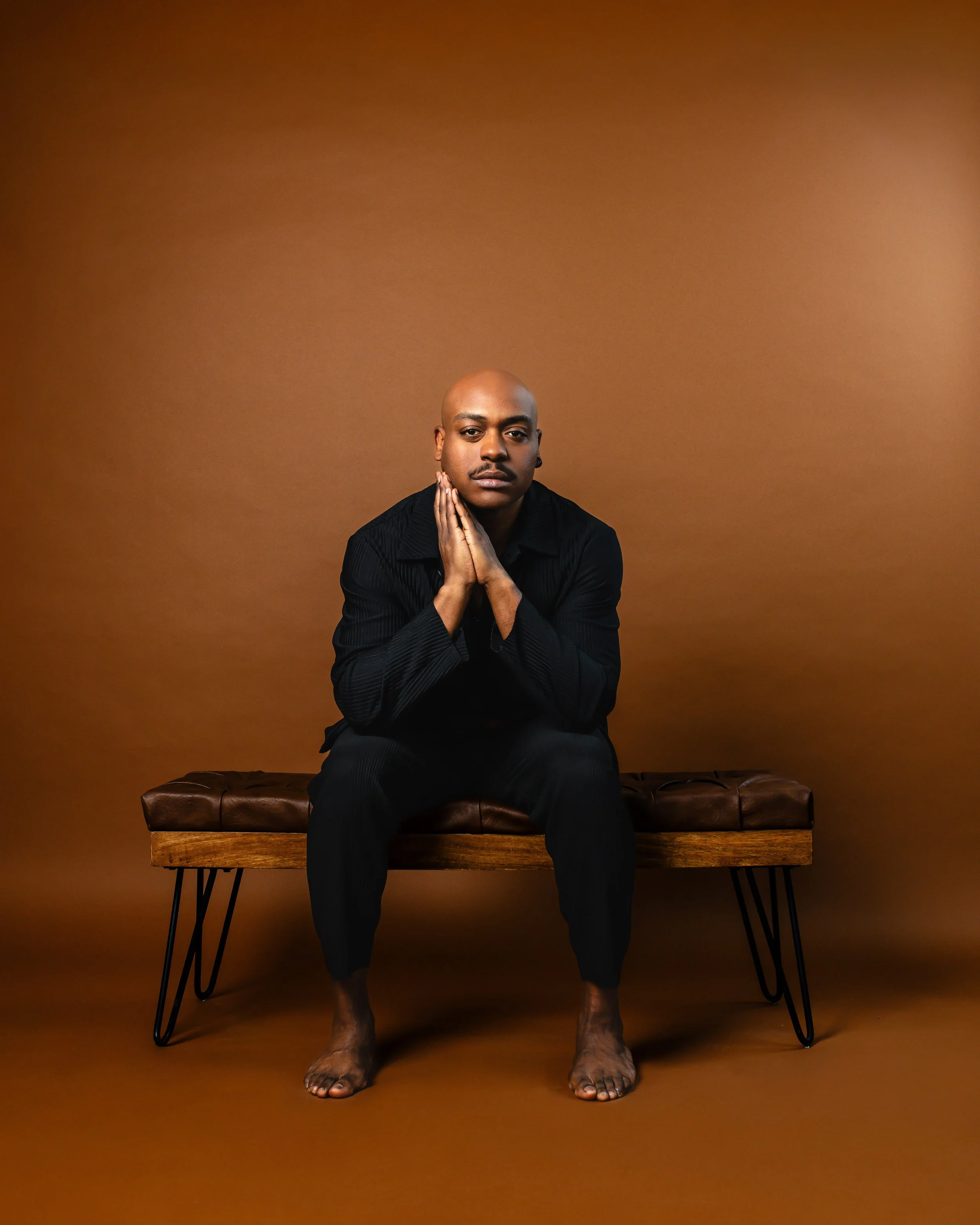 A bald man with dark skin wearing a black outfit sits on a brown leather bench with modern metal hairpin legs; he is barefoot and has his hands pressed against his cheeks, looking contemplative against a plain brown background.