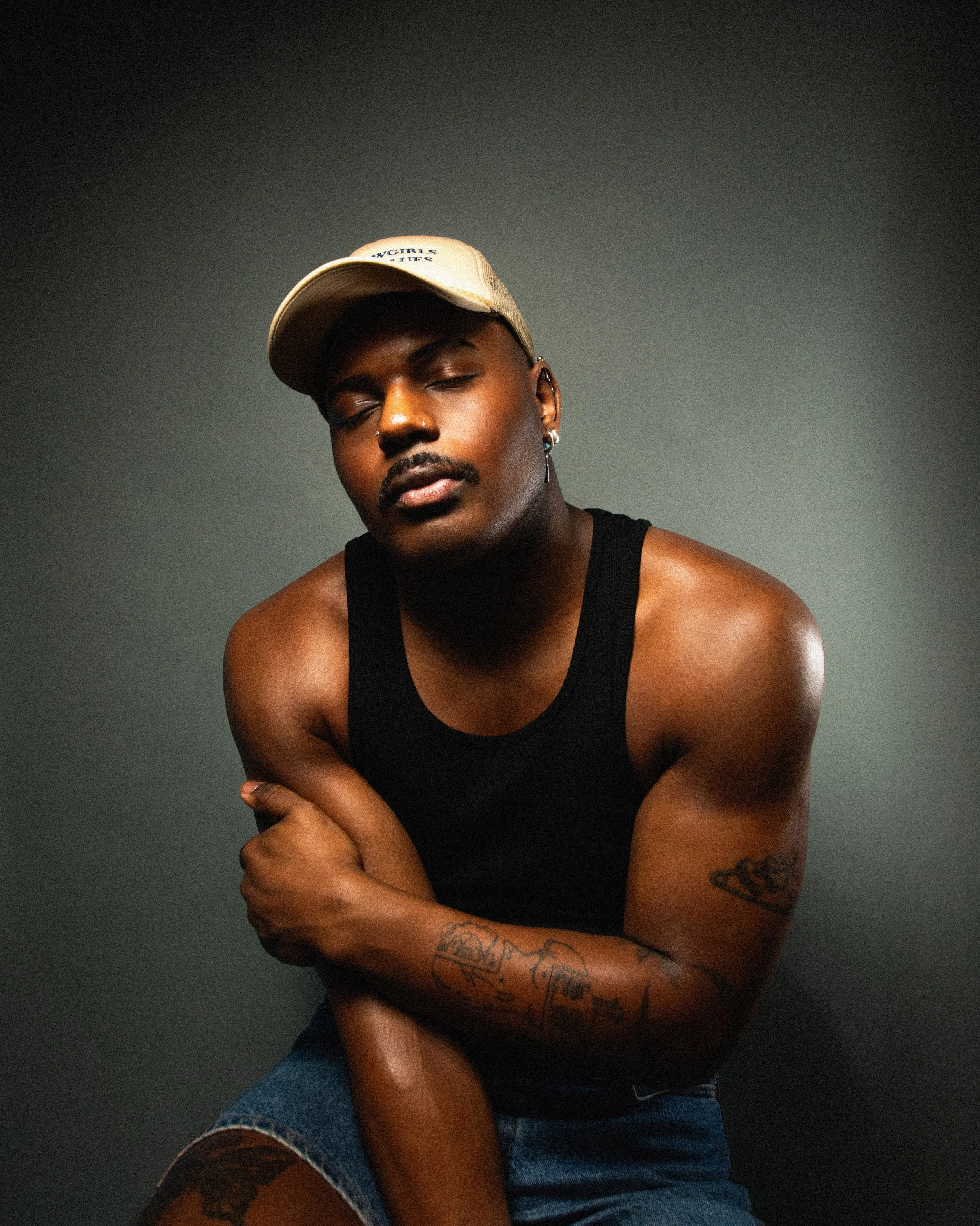 A young man with dark skin and tattoos wearing a beige baseball cap, a black tank top, and jeans, posing against a dark gray background with a relaxed expression.