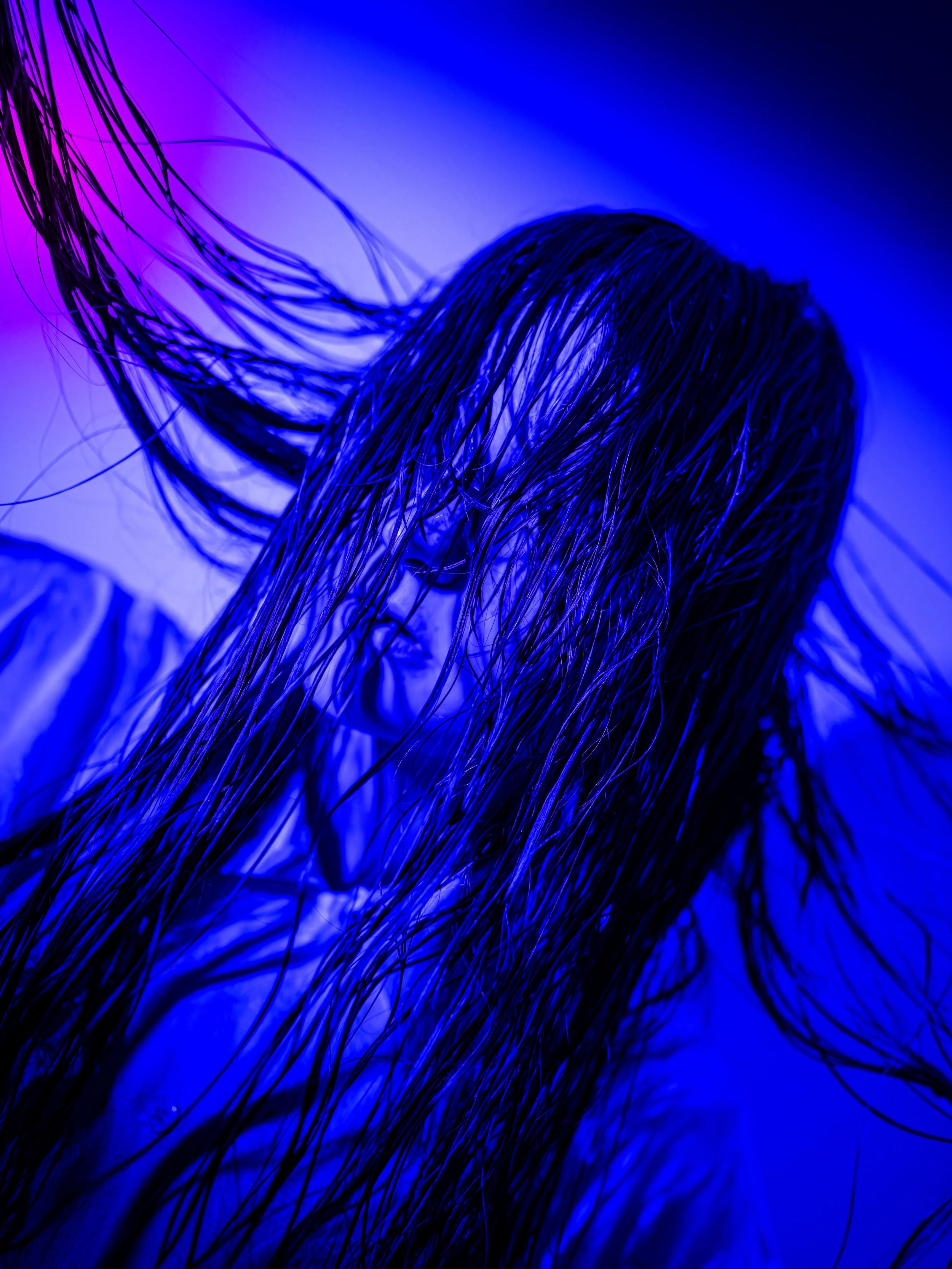 A woman with long, wet hair and a serious expression, illuminated by blue and purple lighting.