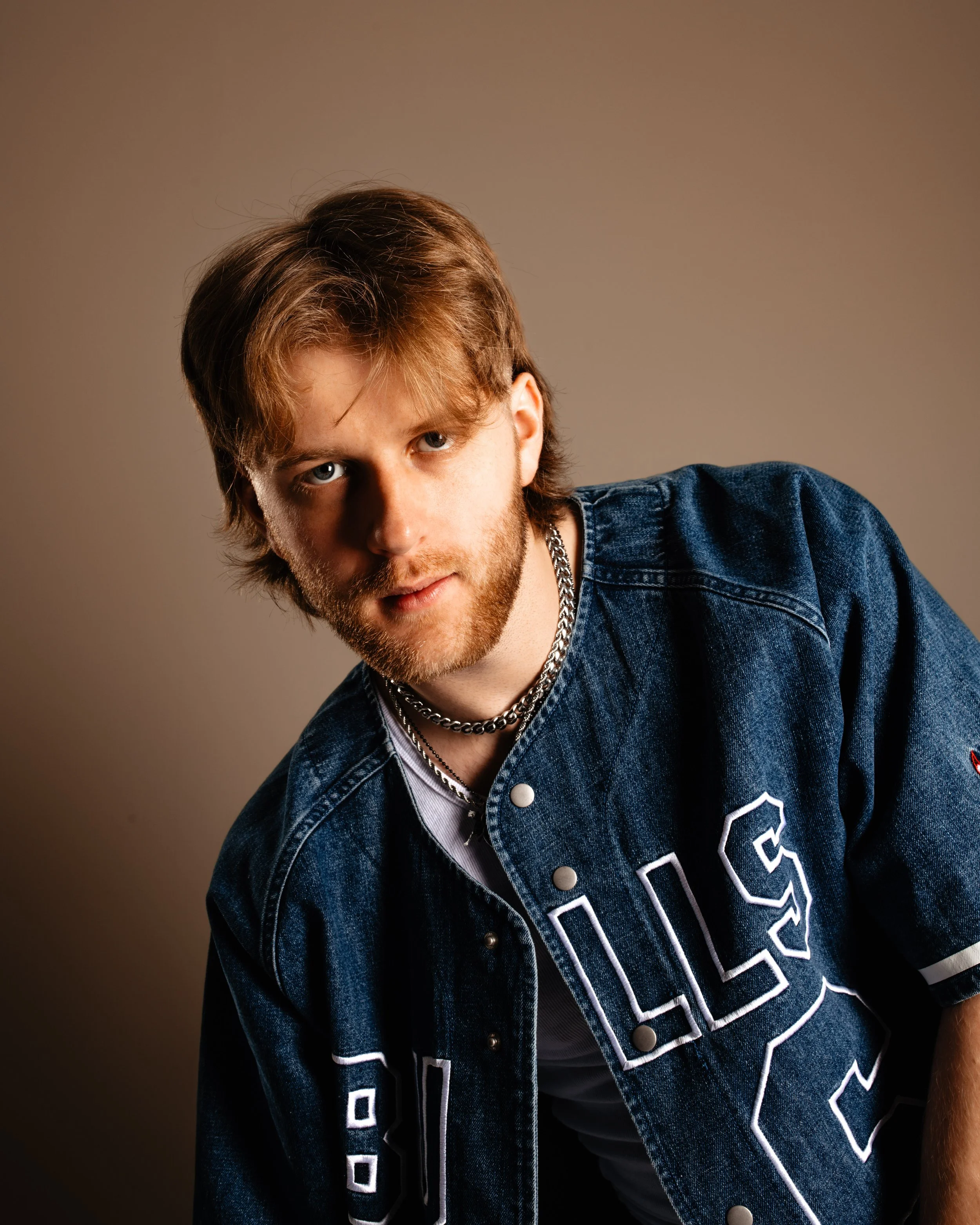 A young man with brown hair and a beard, wearing a denim jacket with letter patches, layered necklaces, and a white T-shirt, posing against a plain beige background.