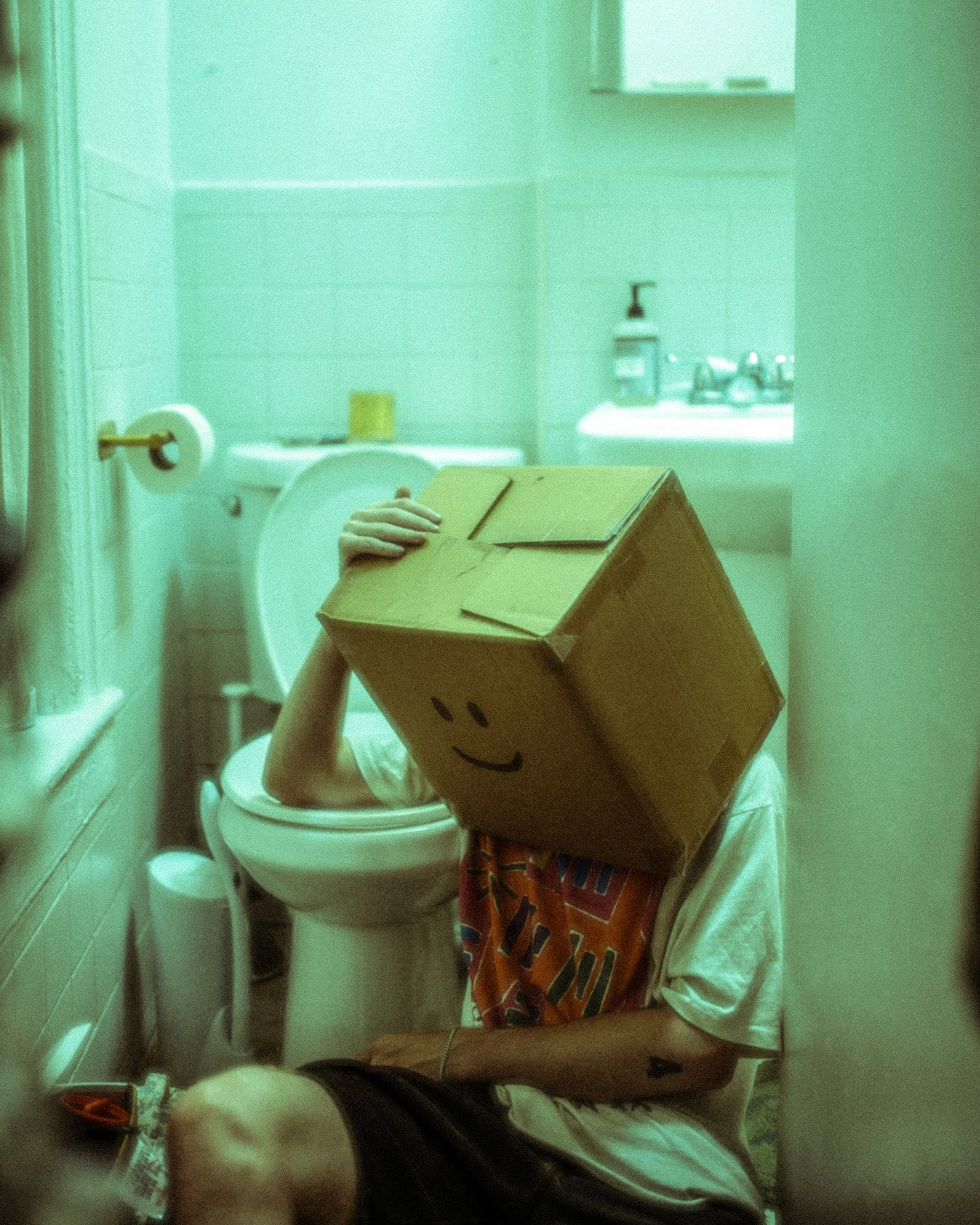 Person sitting on toilet in a bathroom with a cardboard box on their head, drawn with a smiley face, holding it with one hand.
